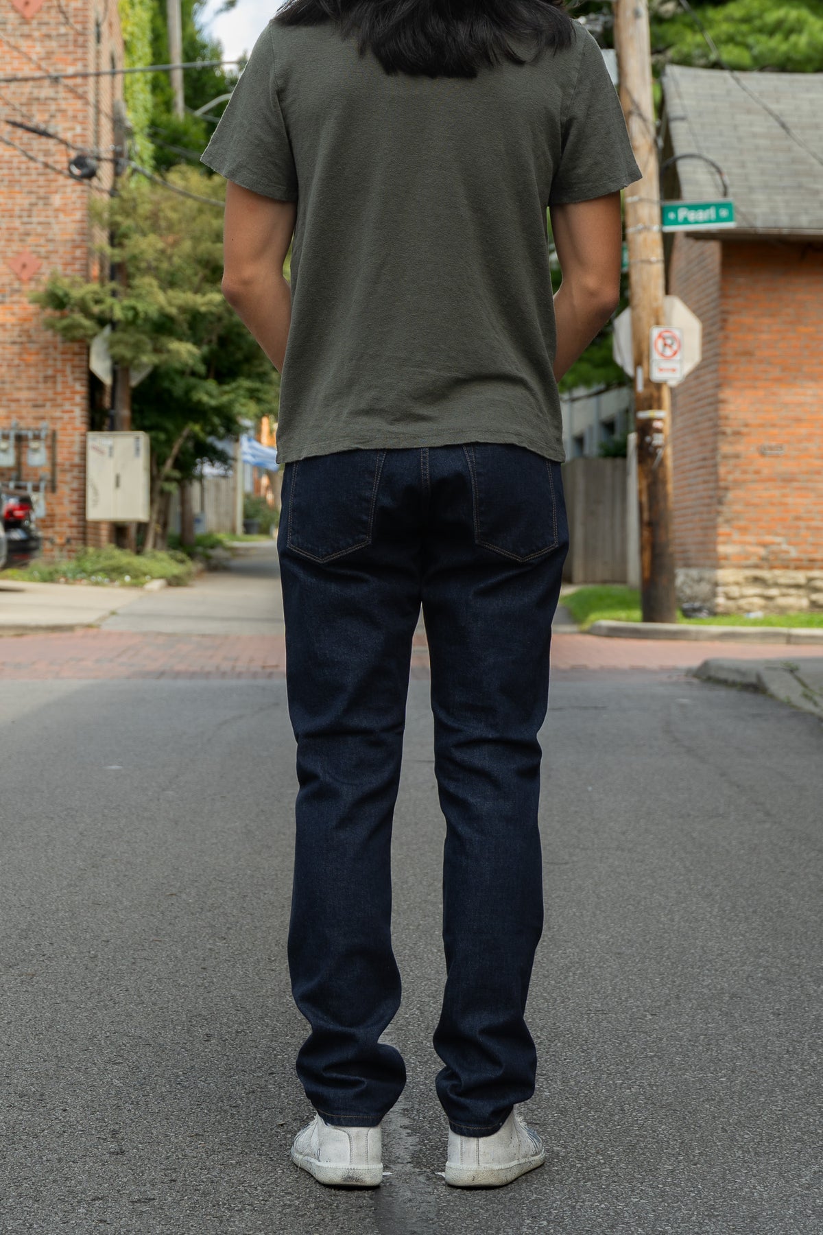 A person standing on a street, viewed from behind, wearing a dark olive green t-shirt, dark blue slim-fit jeans, and white sneakers.
