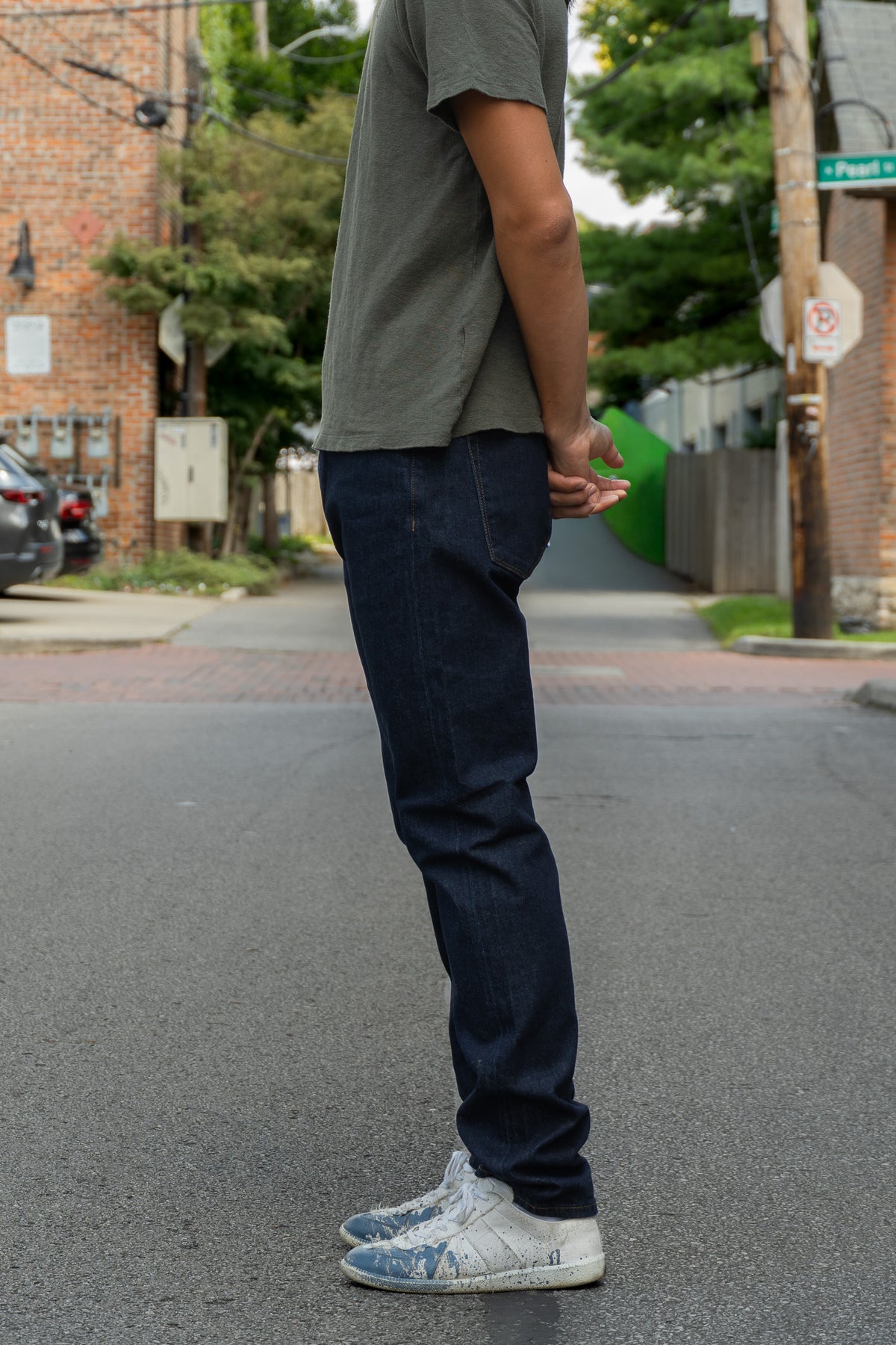 A person standing in the street wearing dark blue jeans, a green t-shirt, and white sneakers, shown from the side.