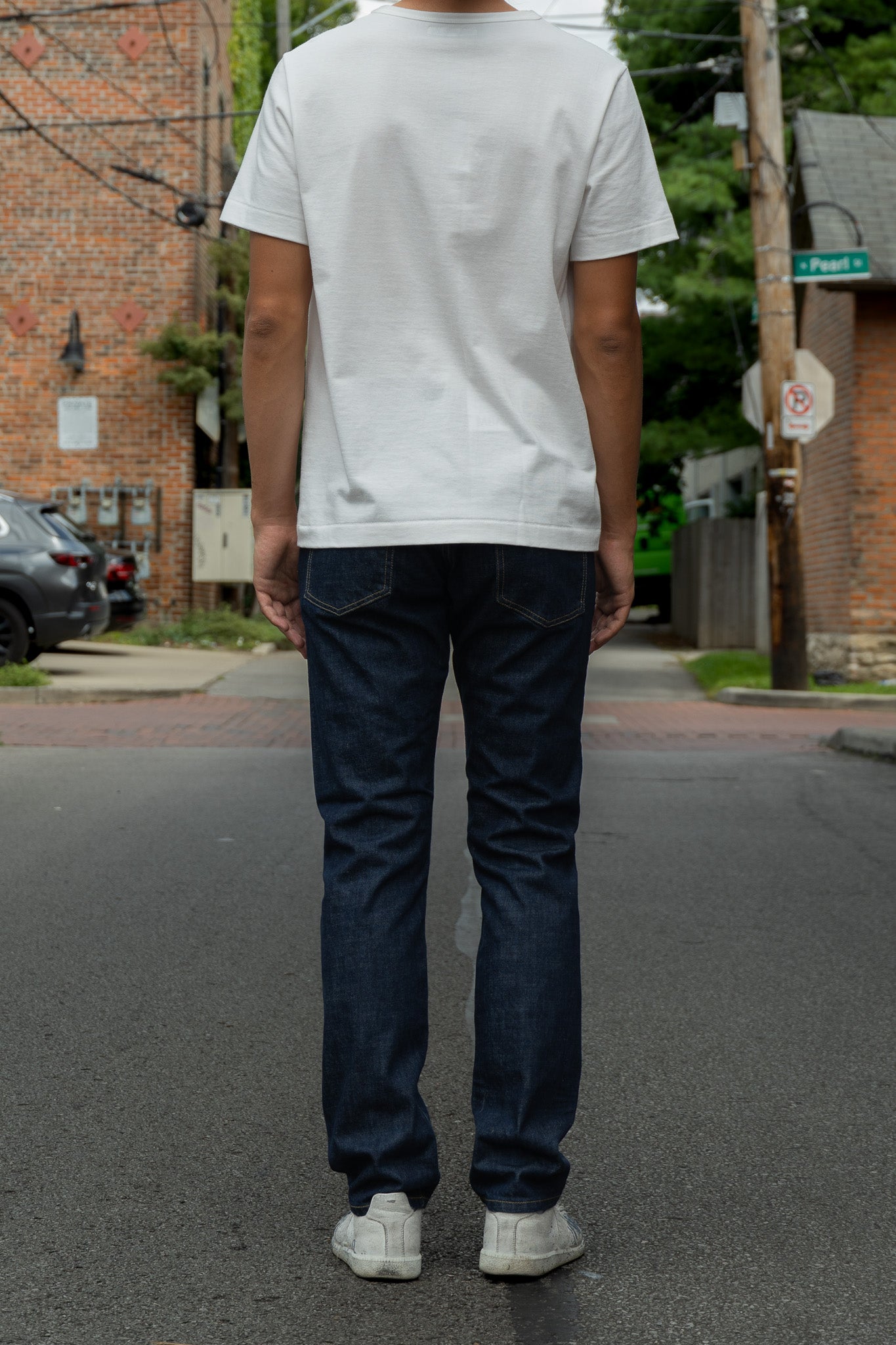 A person standing on a city street, viewed from behind, wearing a plain white short-sleeve T-shirt, dark blue jeans, and white sneakers.