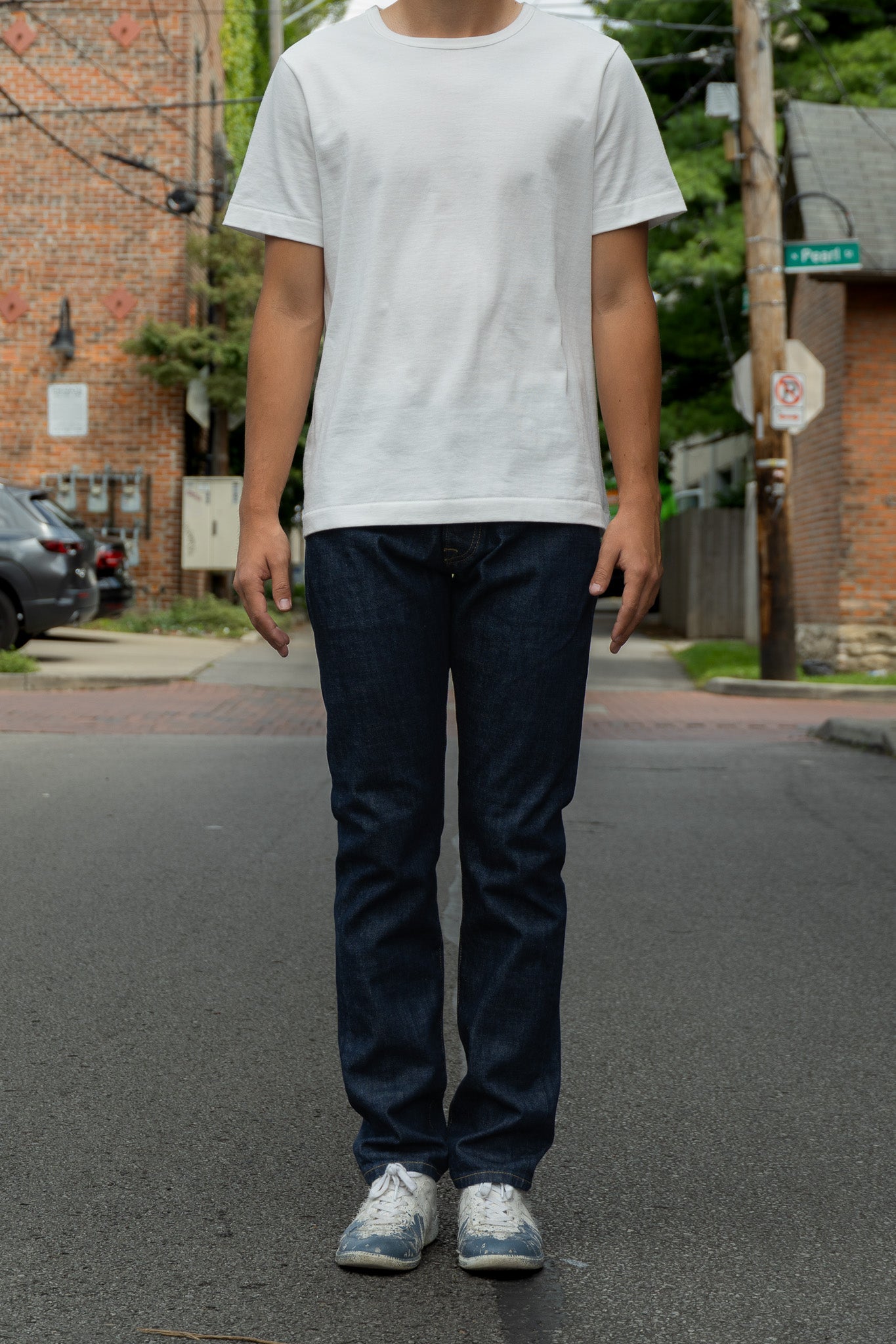A person standing on an outdoor street wearing a plain white short-sleeve crewneck T-shirt, dark blue jeans, and light-colored sneakers.