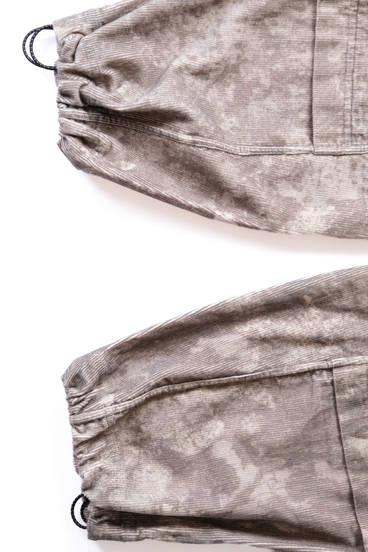 Close-up of a pair of cargo pants with a gray and beige winter camo, corduroy texture, featuring elastic cuffs with black toggles around ankles for adjustability, laid flat on a white background.