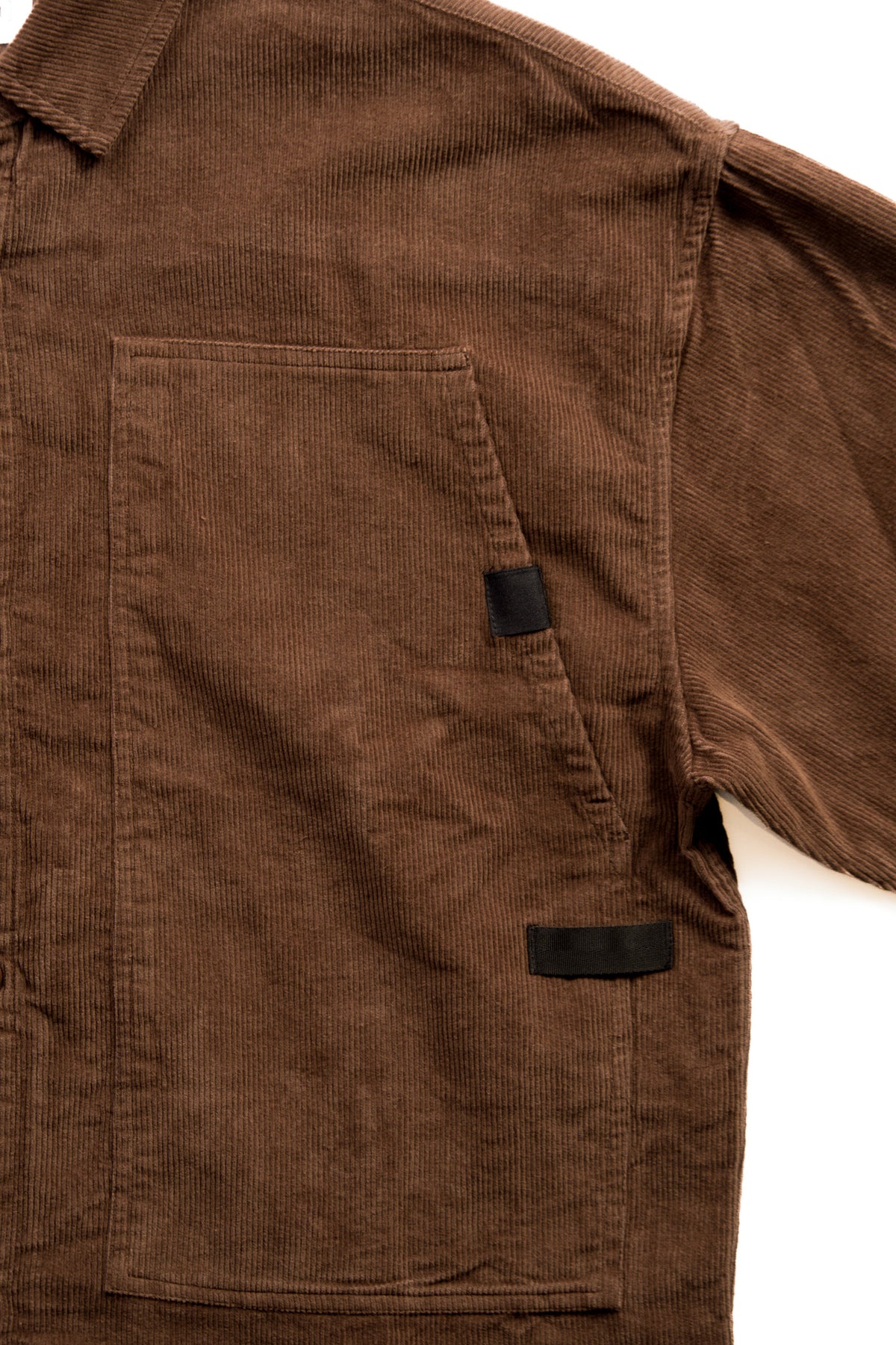 Close-up of a brown corduroy shirt jacket with a large front pocket and two small black fabric tab details, shown on a white background.