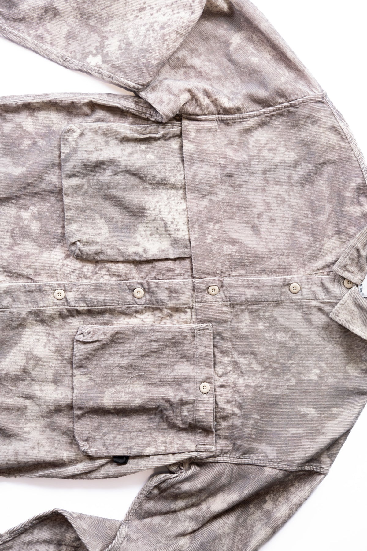 A close-up of a gray and beige winterfied camo corduroy button up showing a pocket, button closure, and textured material on a white background.