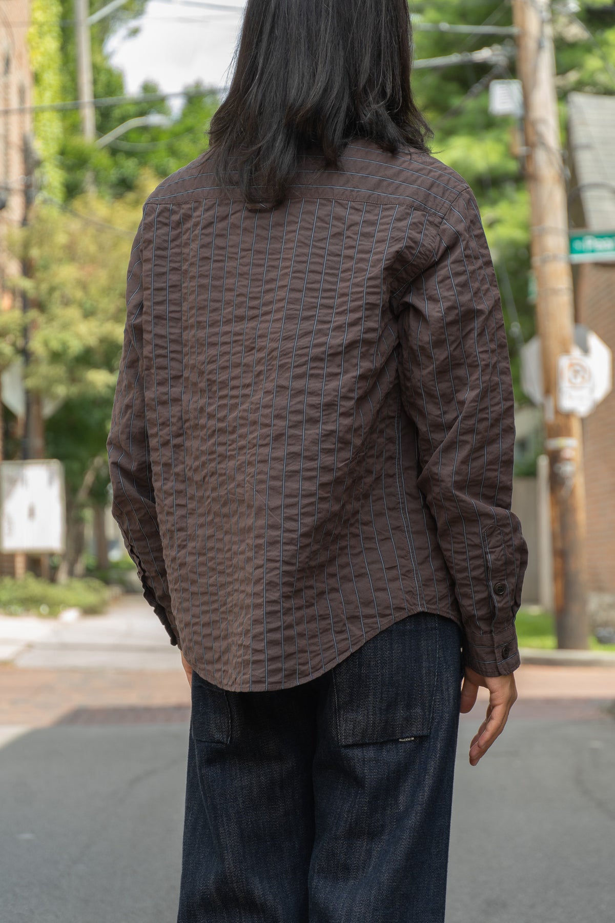 A person viewed from behind wearing a dark brown textured shirt and dark blue jeans, standing on an outdoor street