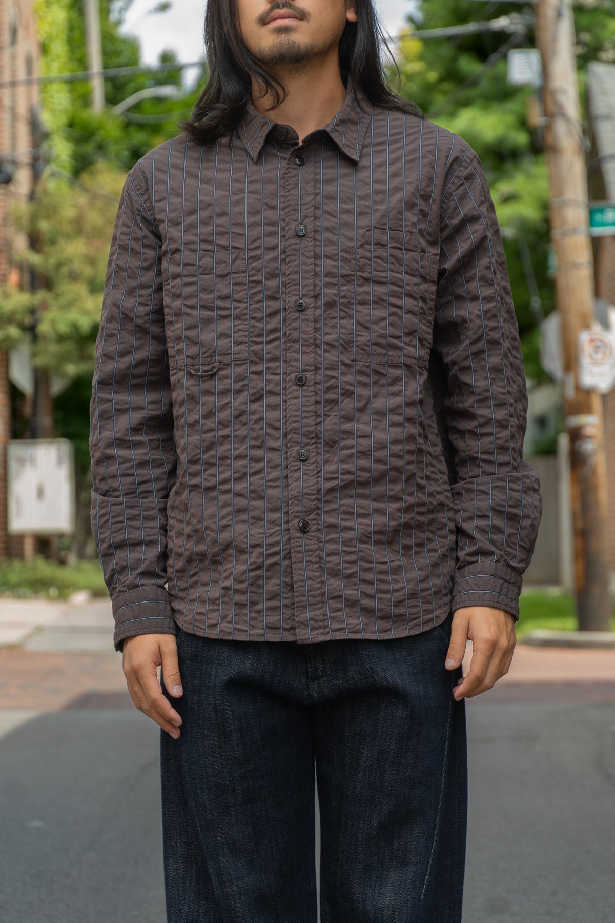 A man wearing a dark brown textured button-up shirt with vertical stripes and dark blue jeans, standing outdoors on a street