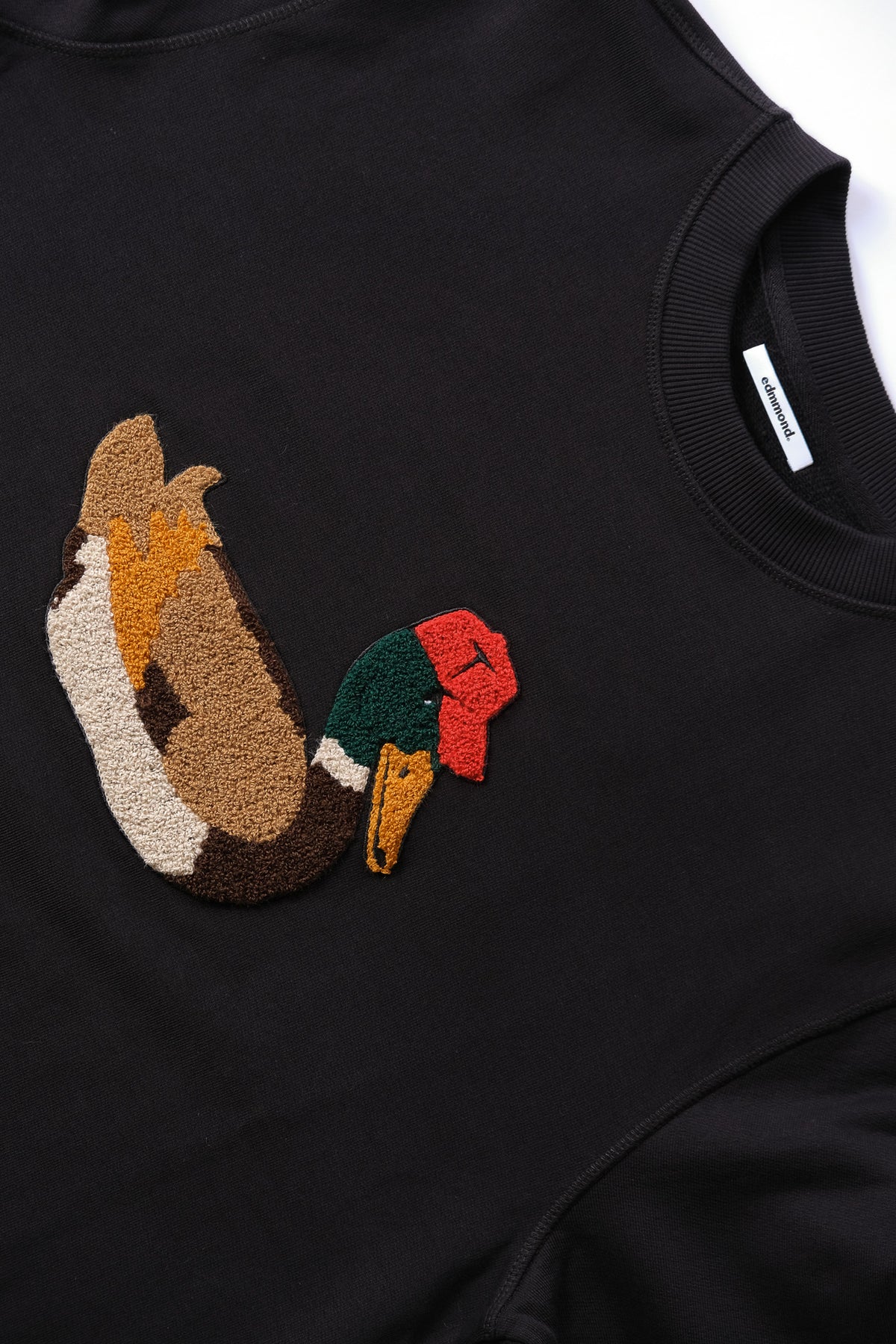 Close-up of a black crewneck sweatshirt featuring a raised, tufted embroidered chest graphic of a duck.