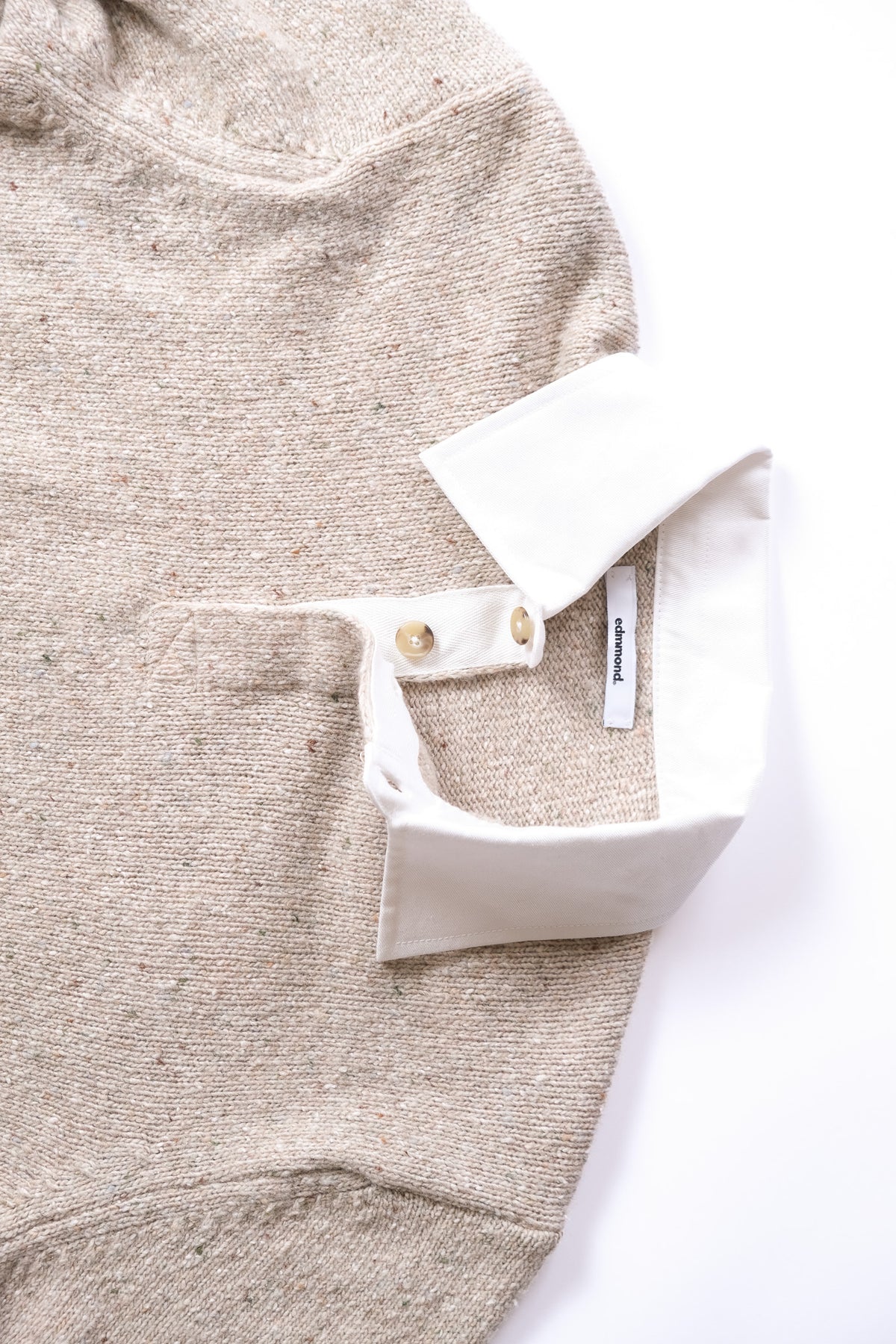 Close-up of a taupe sweater with a white collar turned up, showing two small buttons and a white interior label on a white background.