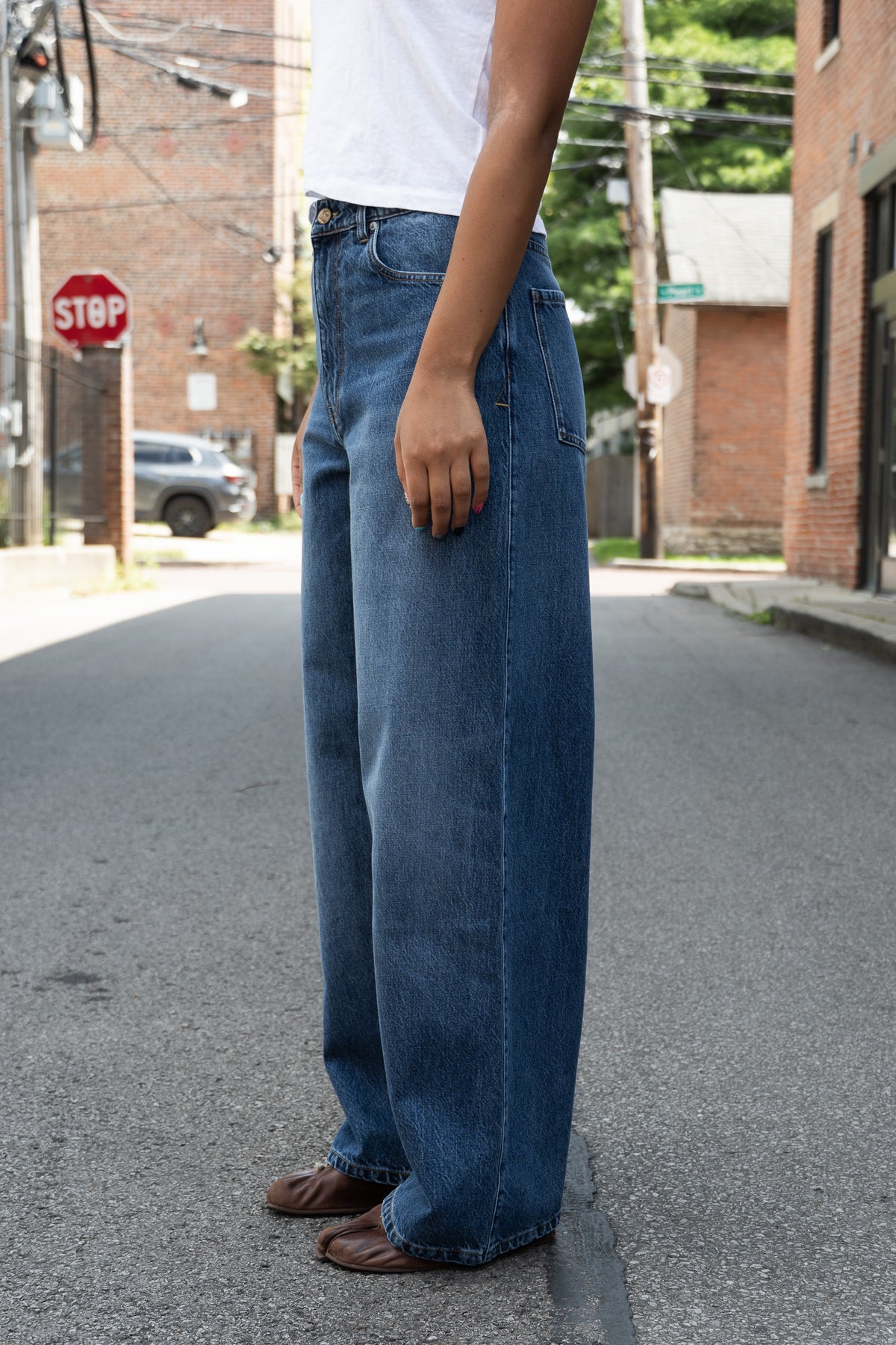 A person standing in the street wearing high-waisted, wide-leg blue denim jeans, a white t-shirt, and brown leather shoes.