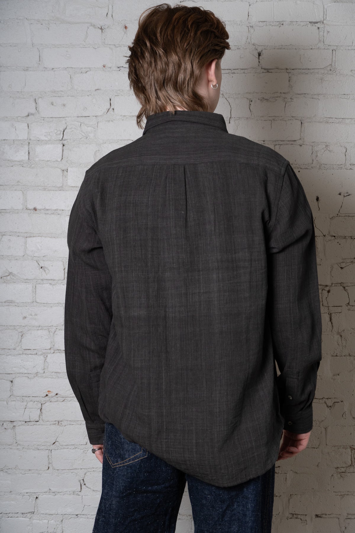 a man wearing a dark charcoal button up shirt photographed from behind on a white brick wall.