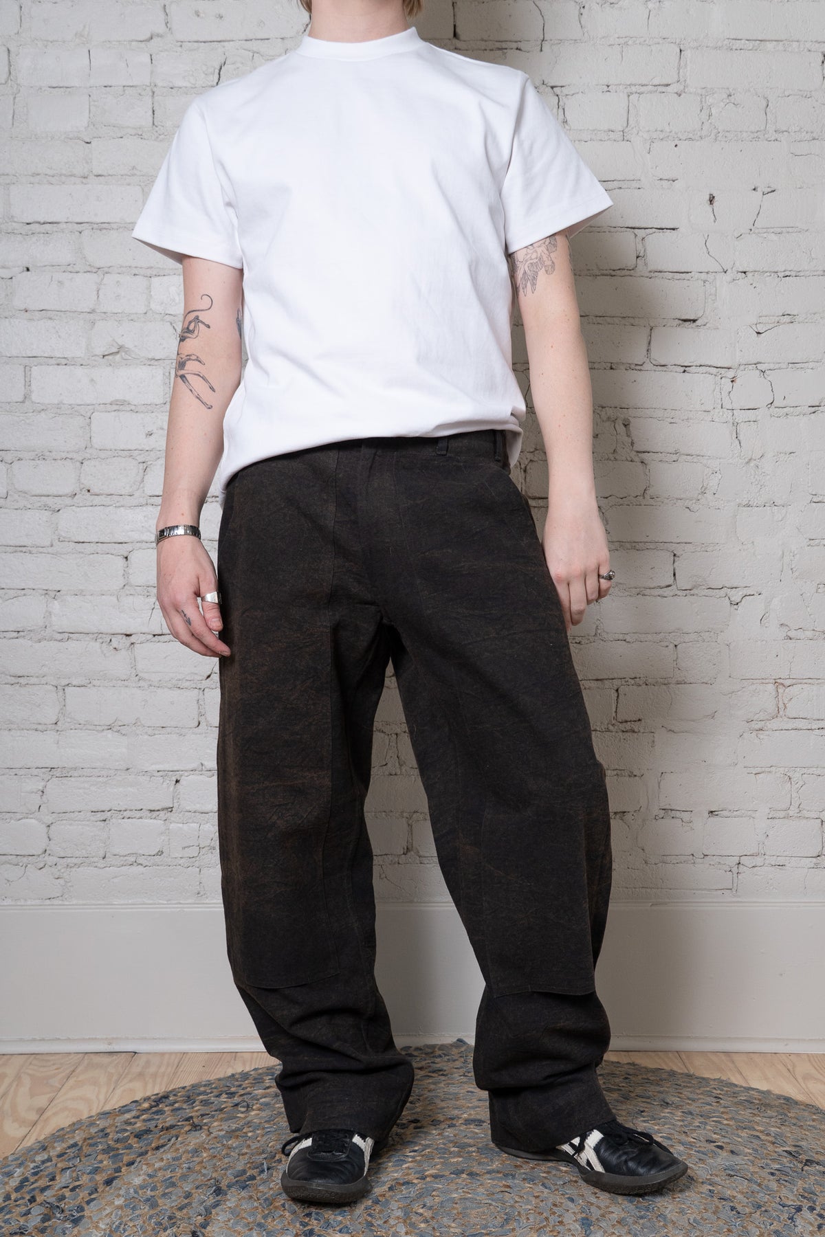 A person standing against a white brick wall wearing a plain white short-sleeve T-shirt, loose iron colored pants, and black sneakers with white stripes.