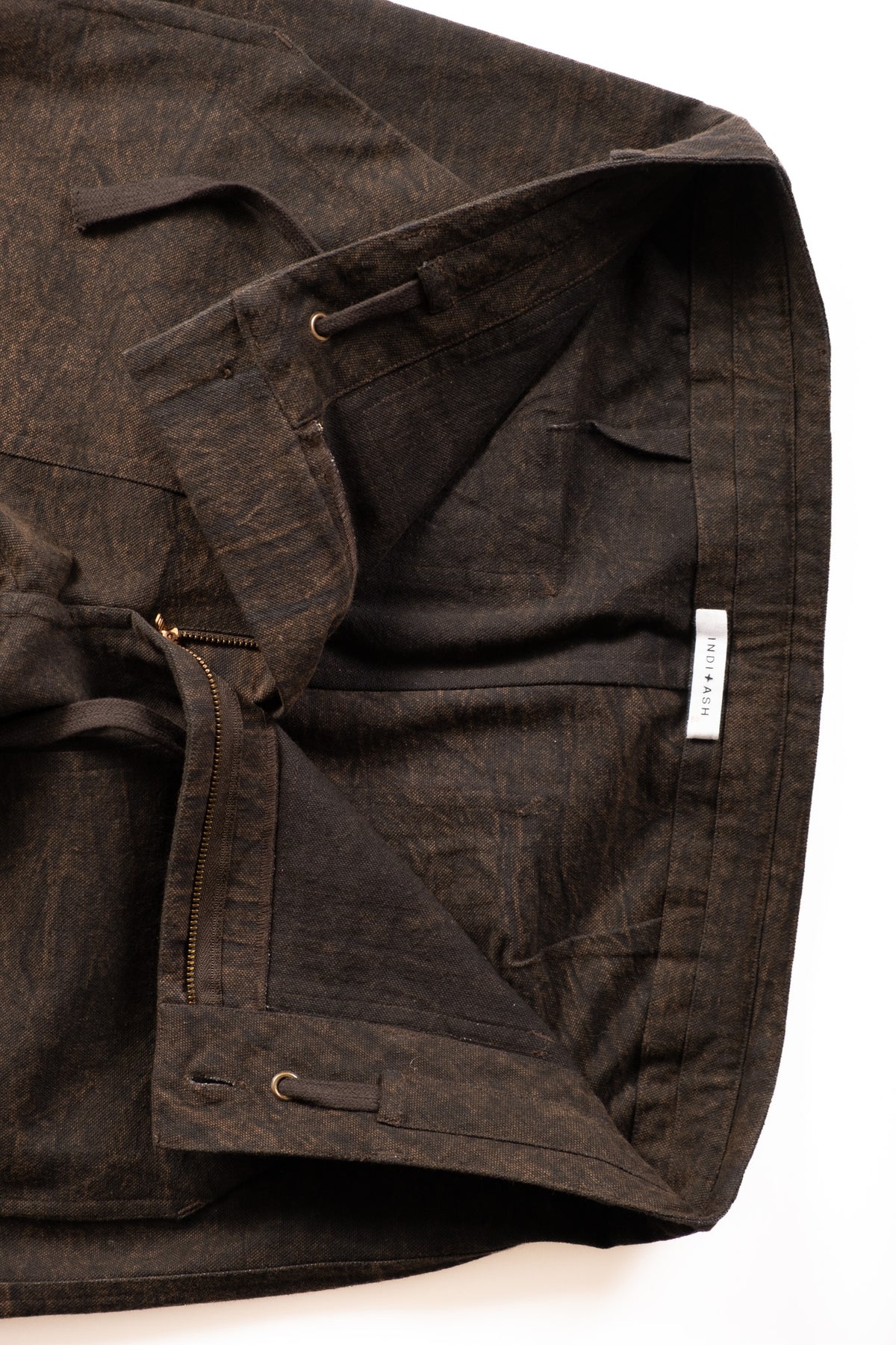 A close-up of a dark brown textured pant laid flat on a white background, showing a zippered pocket, drawstring detail, and a small white brand tag.