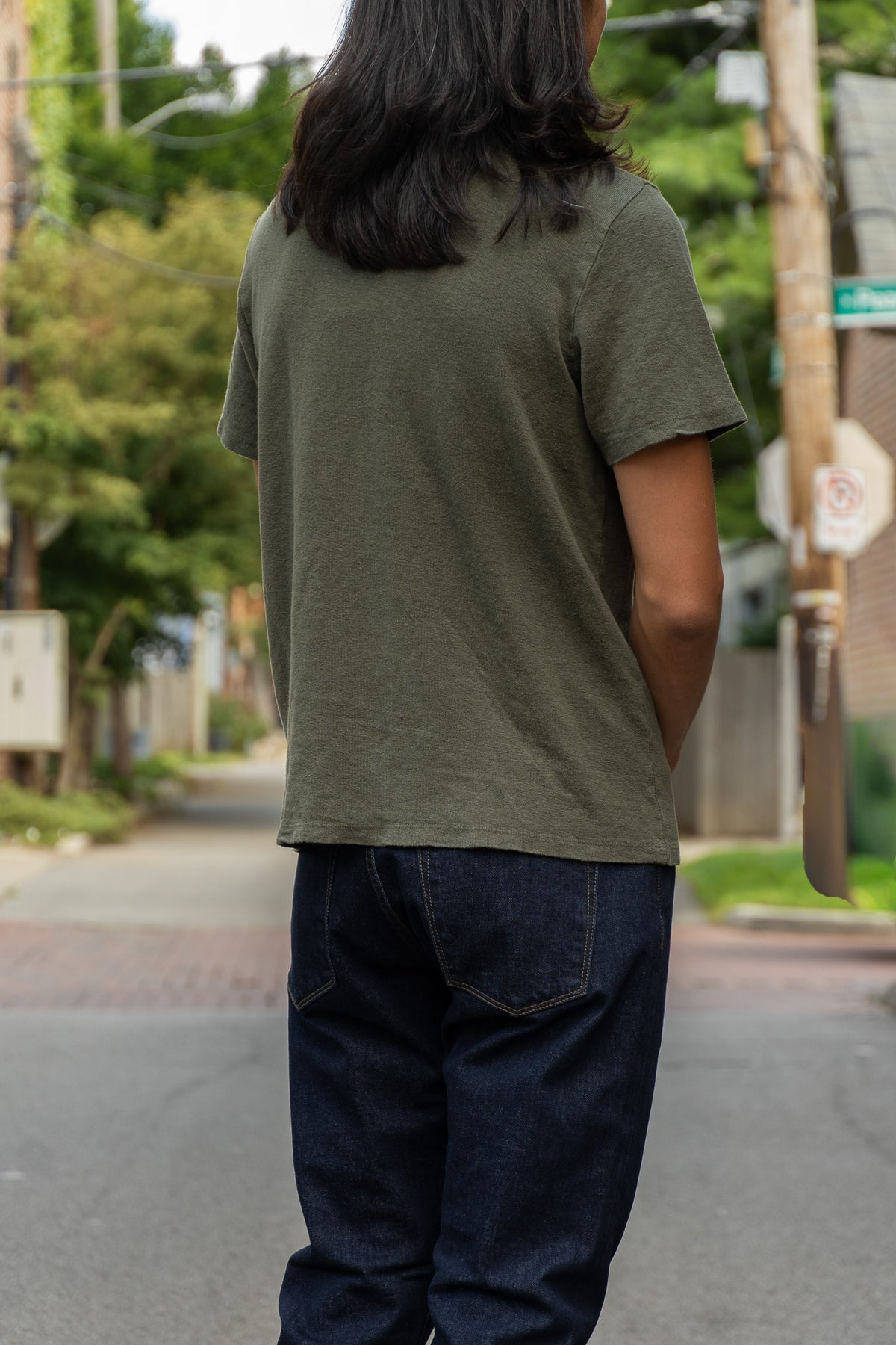 A person viewed from behind wearing an olive green short-sleeve t-shirt and dark blue jeans, standing on a street