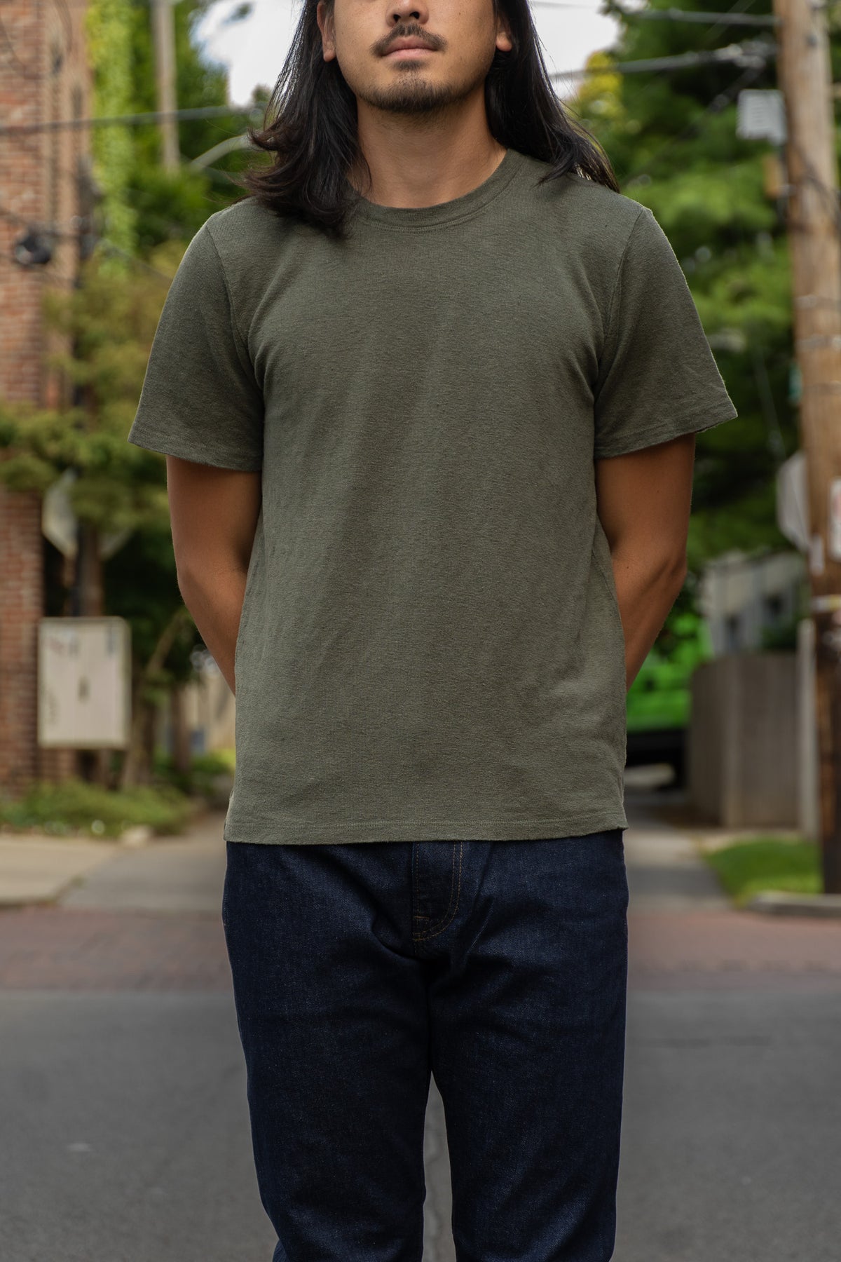 A man wearing an olive green crewneck t-shirt and dark blue jeans, standing on an outdoor street