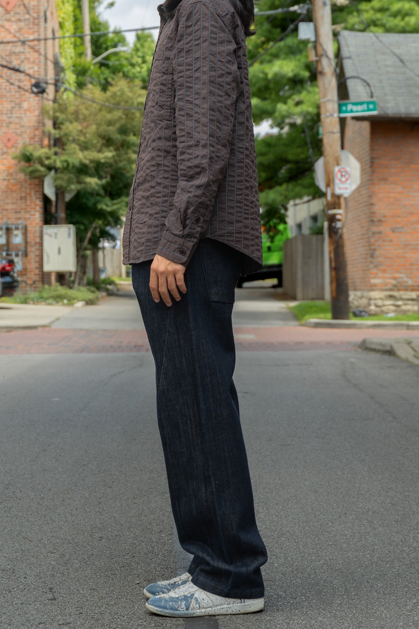 A person standing from the side outdoors on a street wearing a dark brown seersucker shirt, dark blue jeans, and white sneakers.