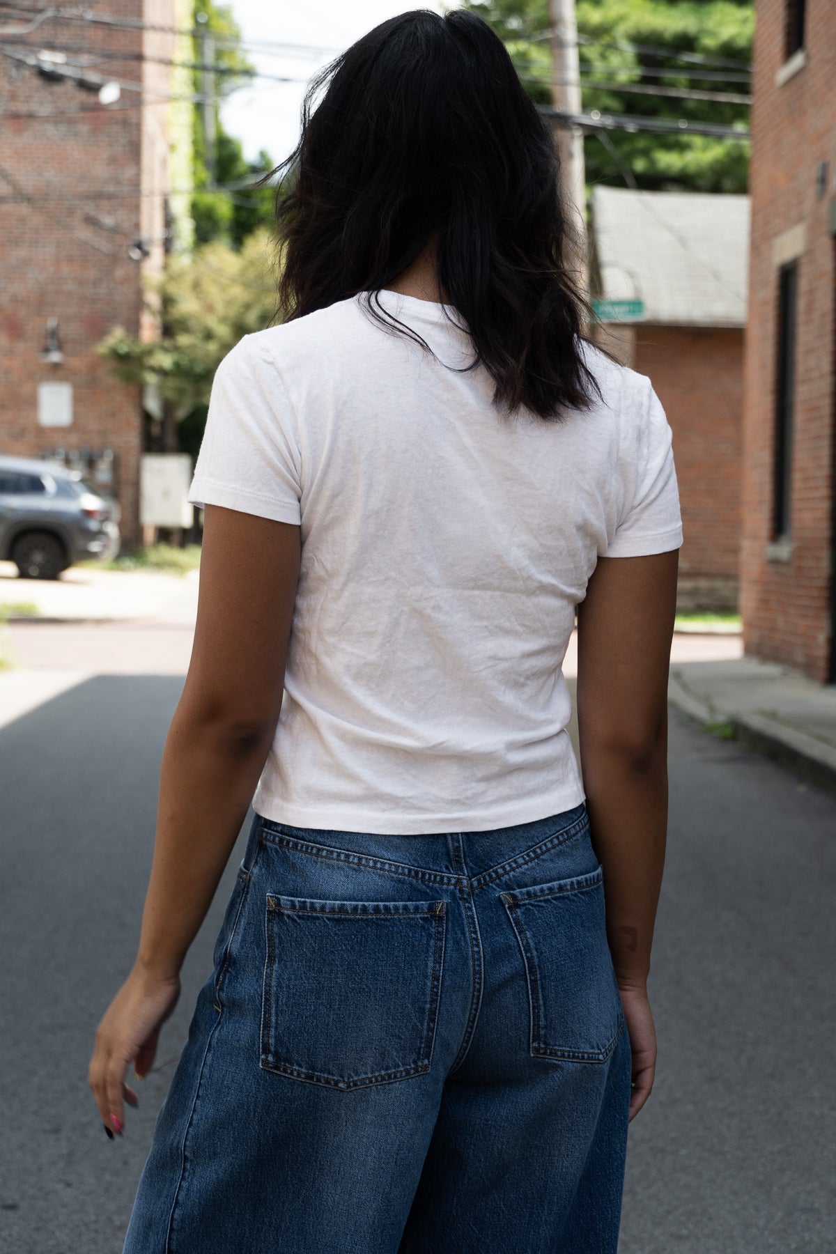 A person viewed from behind wearing a fitted white short-sleeve T-shirt and blue jeans, standing in the street.