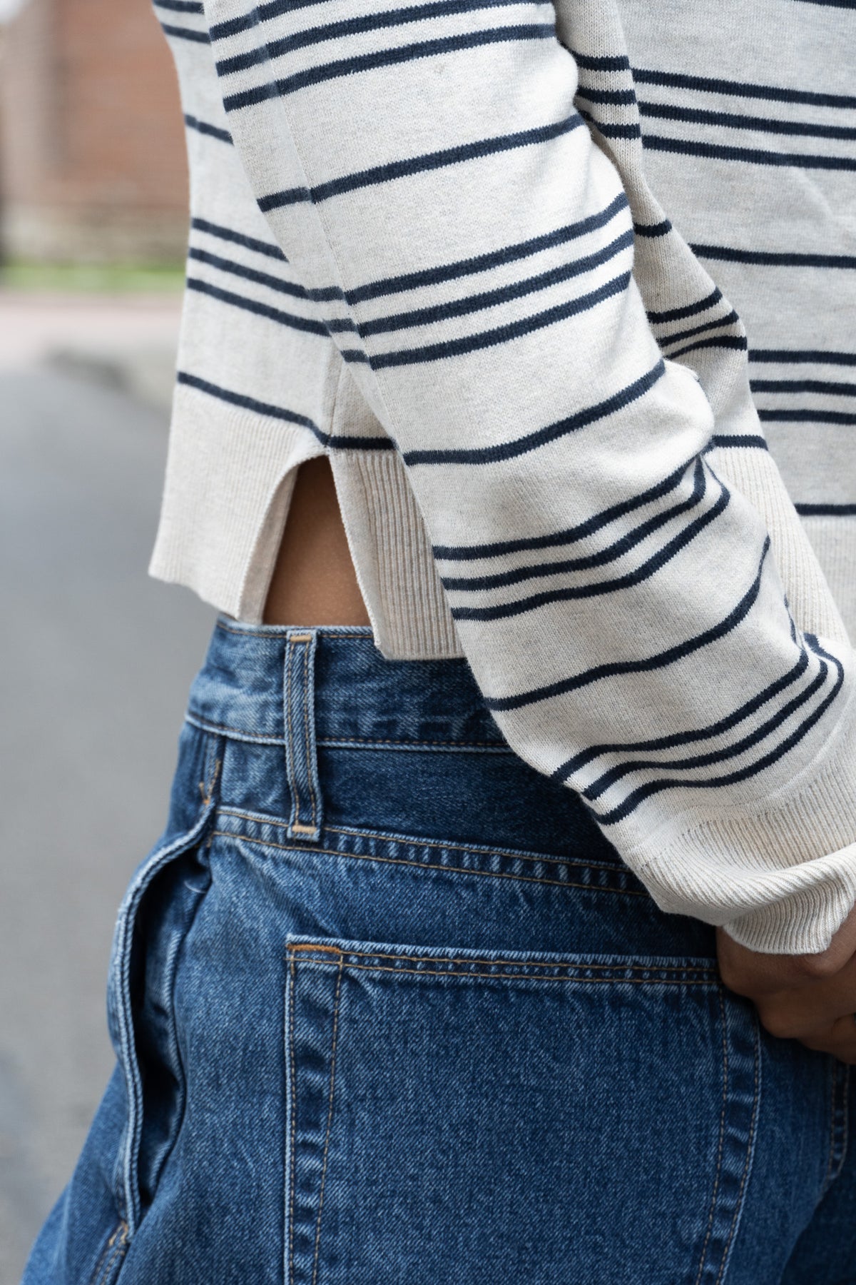 Close-up of a person wearing a cream knit sweater with navy horizontal stripes and a side slit hem, paired with blue jeans.