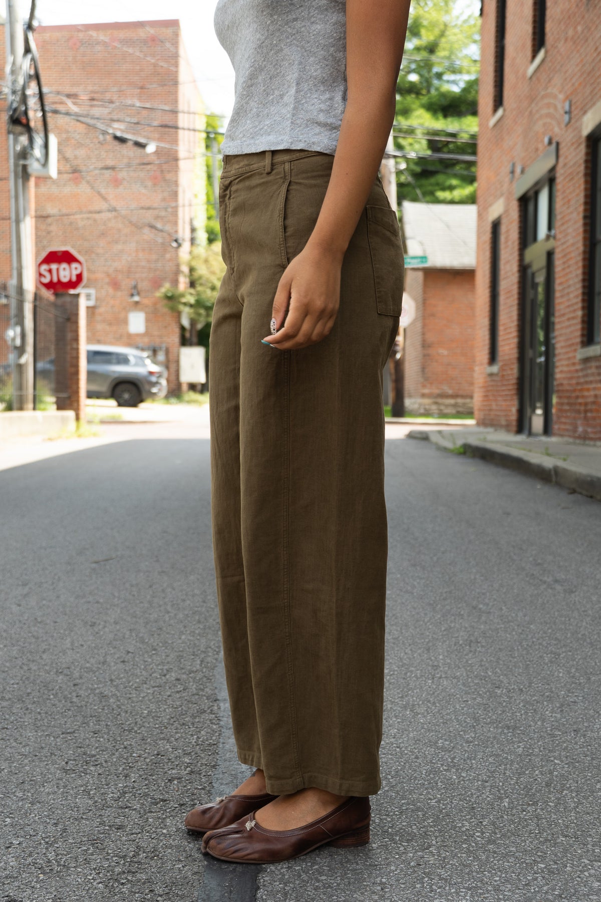 A person standing on an urban street wearing wide-leg olive brown trousers, a light gray sleeveless top, and brown leather flats.