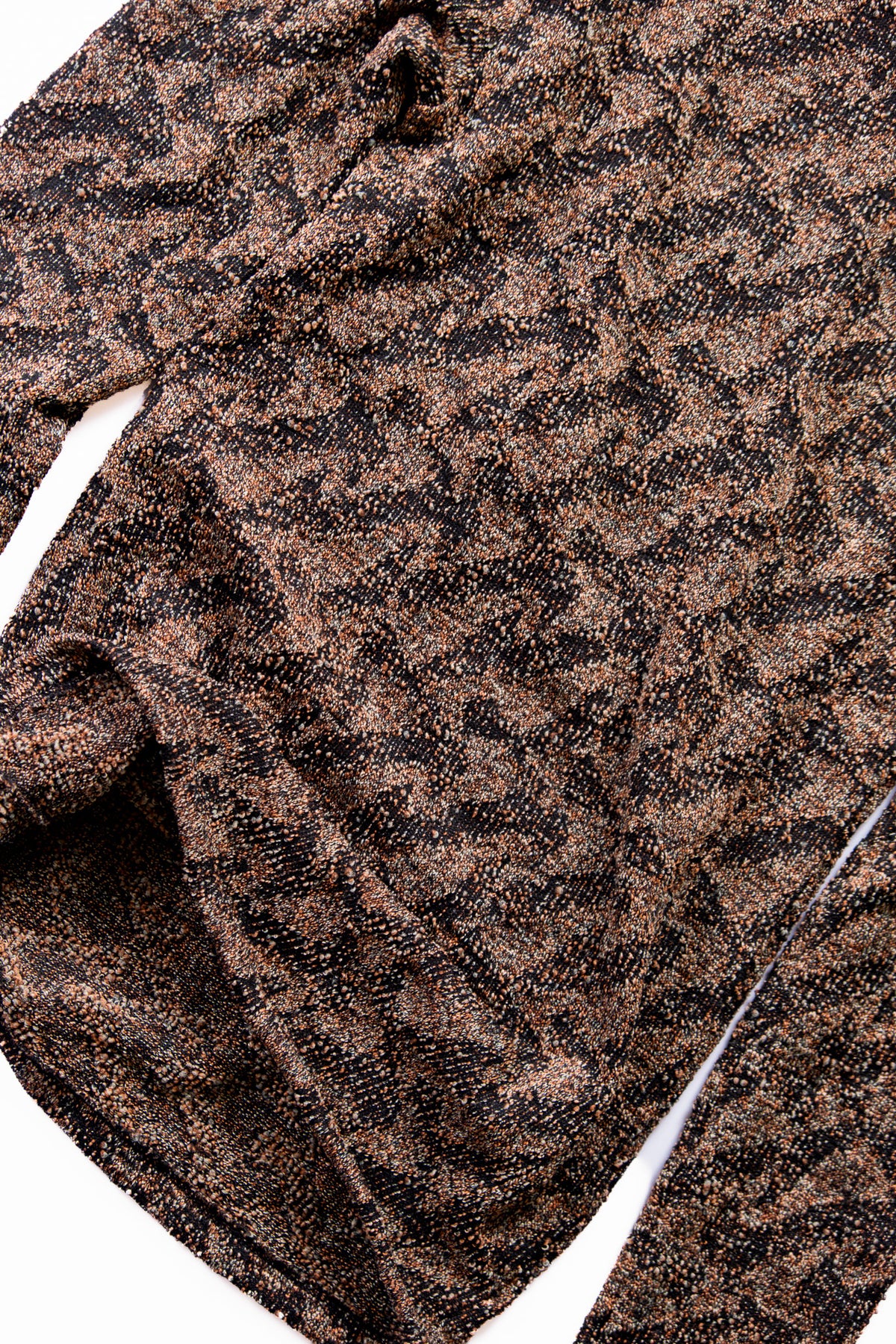Close-up of a textured knit fabric with a mottled brown and black pattern, showing the weave and a portion of a long sleeve.