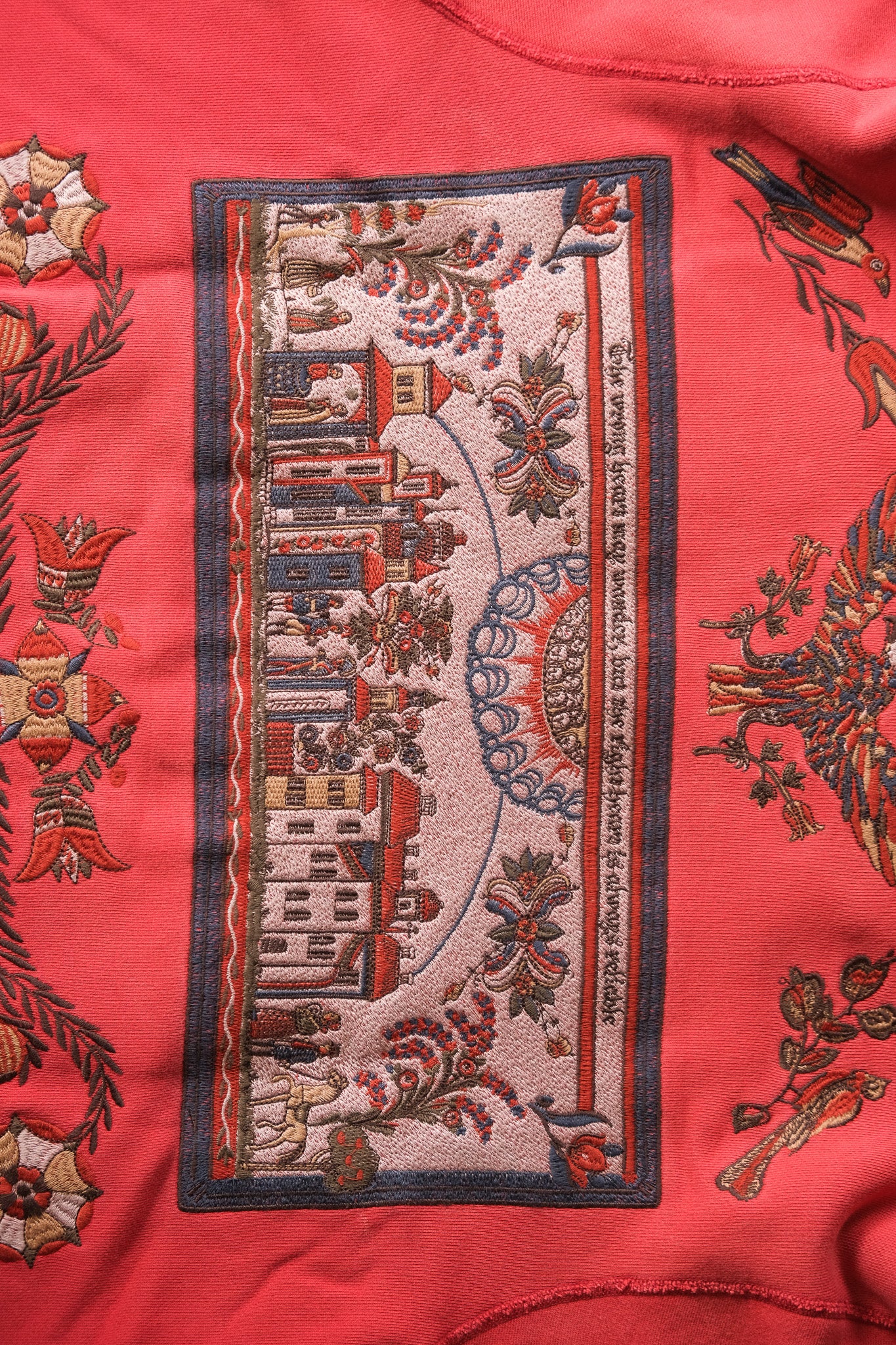 A close-up of a faded red pullover featuring a central rectangular embroidered patch with a vintage cityscape and botanical motifs in muted red, beige, and blue-gray tones.