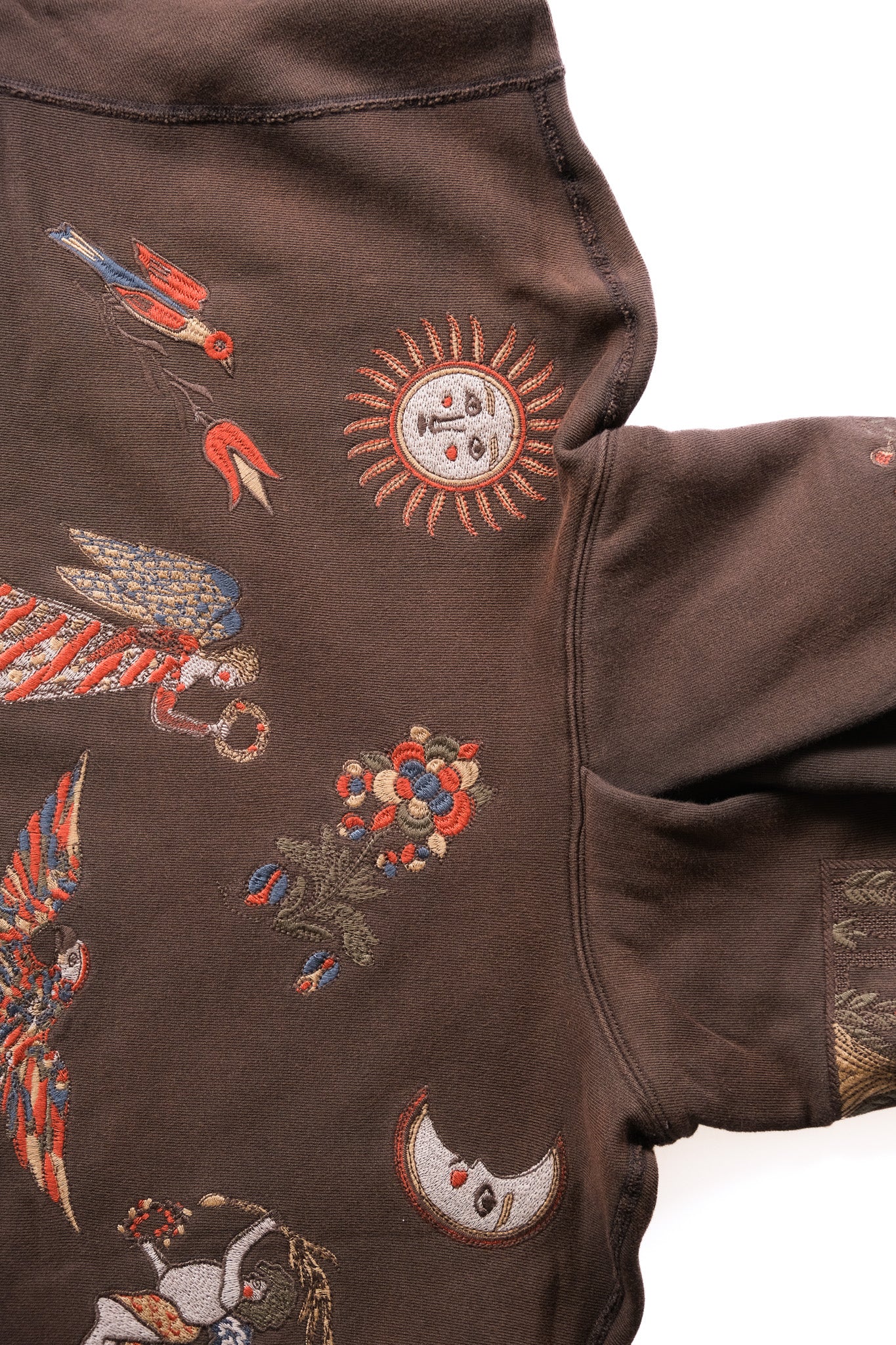 A close-up of a dark brown sweatshirt featuring colorful embroidered motifs including birds, a sun, a crescent moon, and floral patterns.