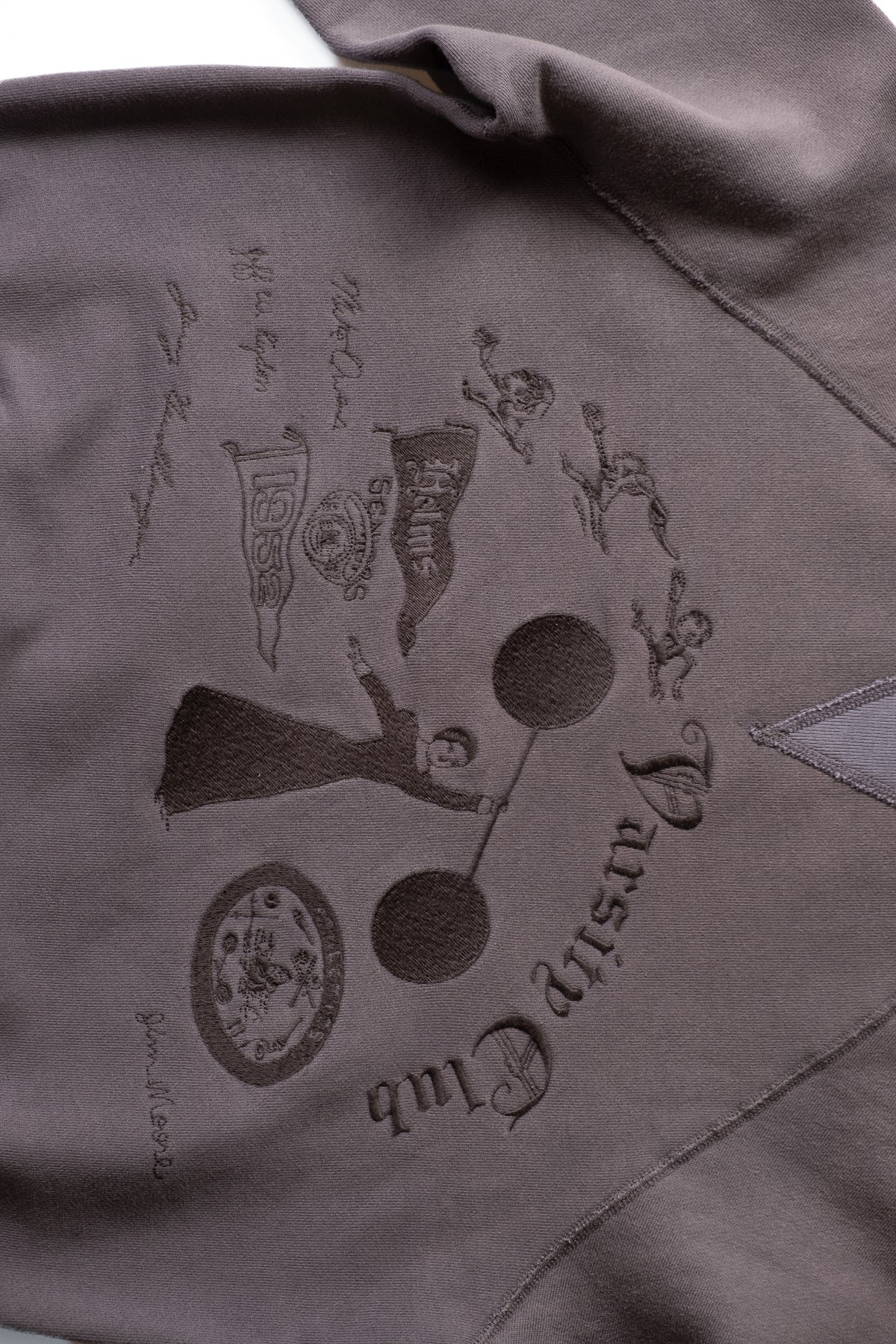 Close-up of a grey crewneck featuring an embroidered graphic of a woman holding a dumbbell, with the words 'Varsity Club'