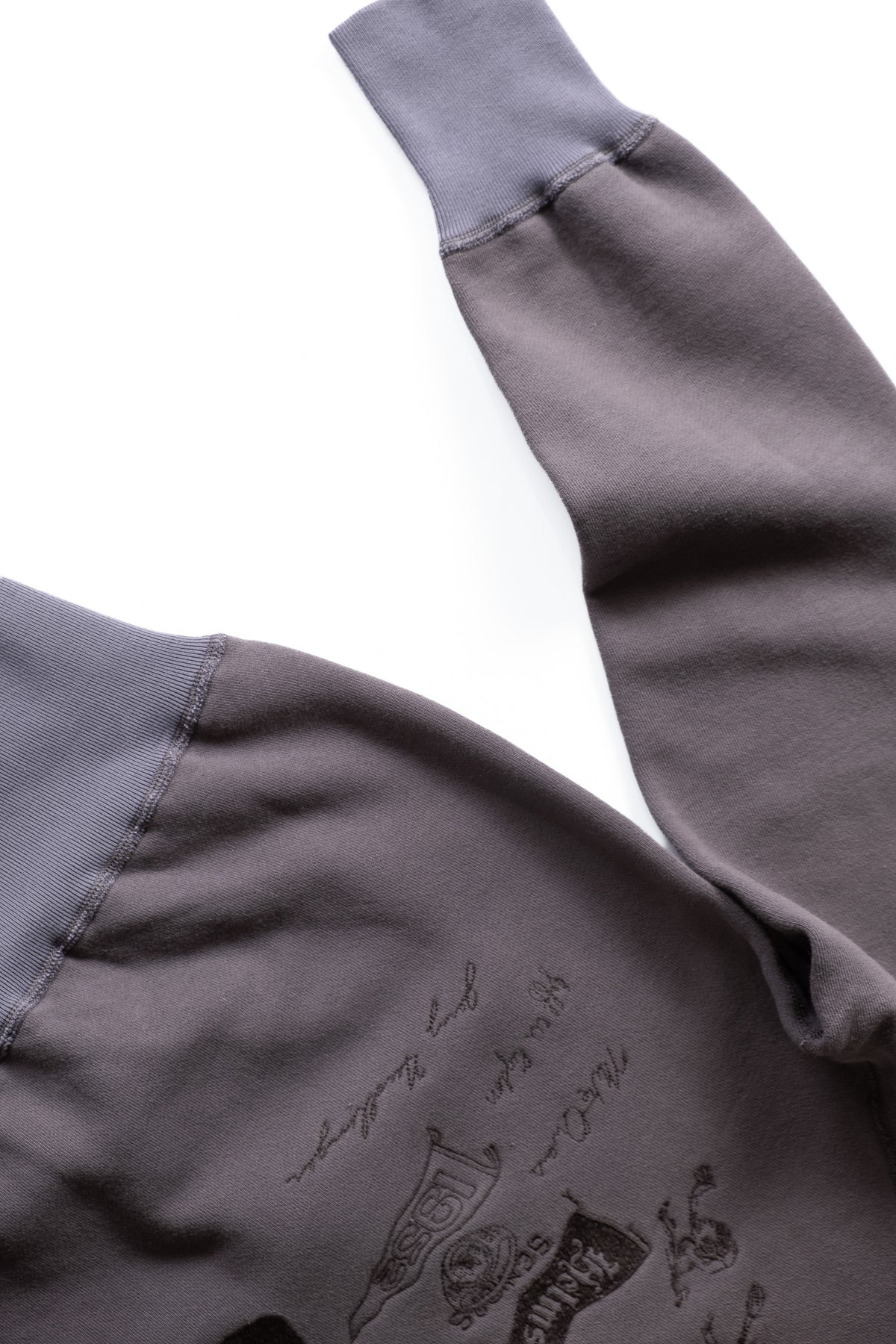 Close-up of a dark gray sweatshirt sleeve with a black embroidered graphic print and script text on the chest, laid flat on a white background.