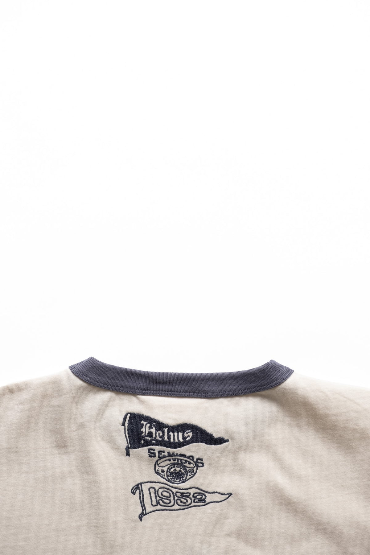 Close-up of the back collar and upper back of a light beige ringer T-shirt with a dark navy ribbed crew neck and printed navy graphics on the back.