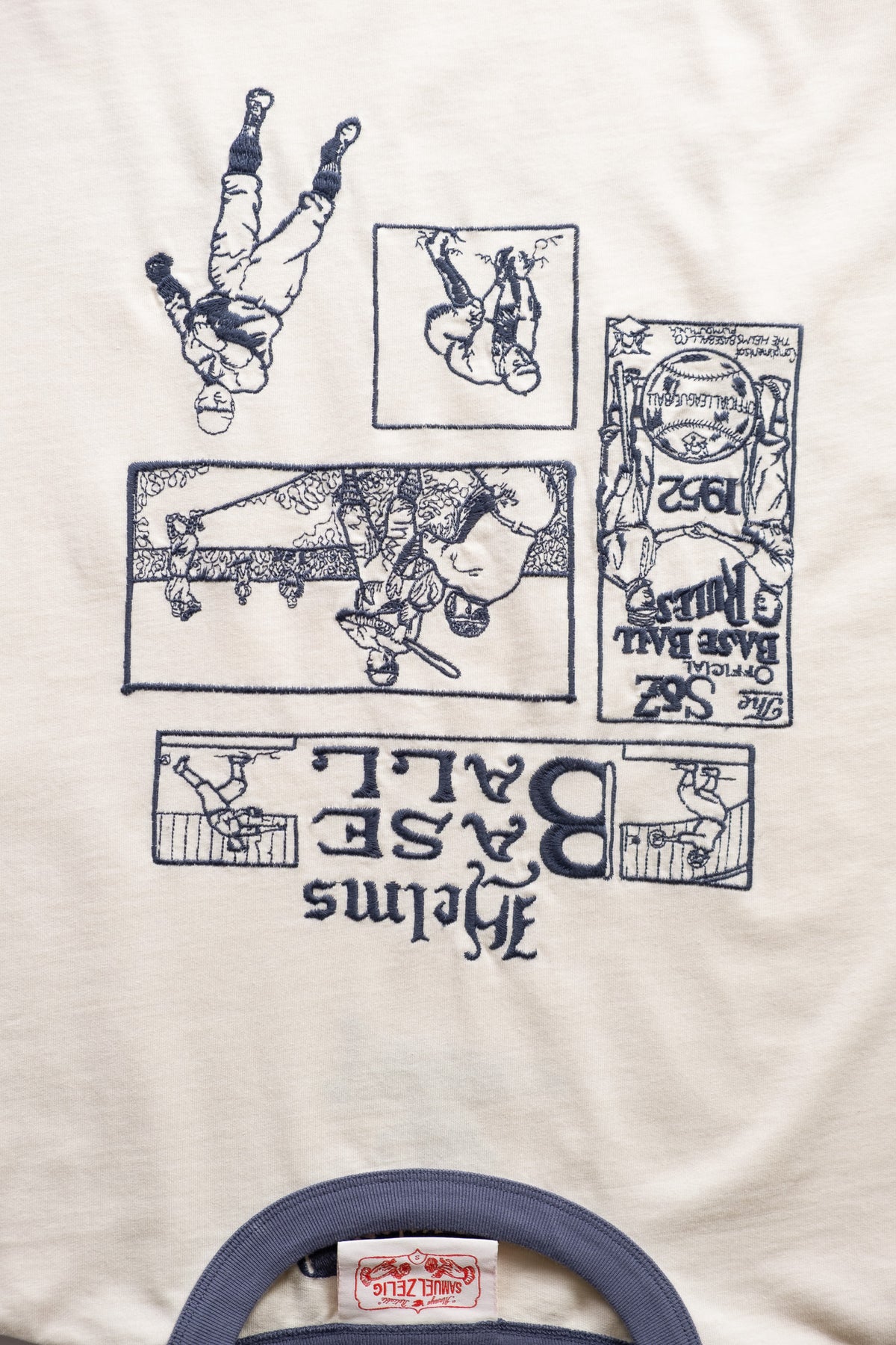 A close-up of a cream-colored t-shirt featuring a blue emboridered graphic print of baseball players and the text 'Helms Base Ball'. A blue collar trim.