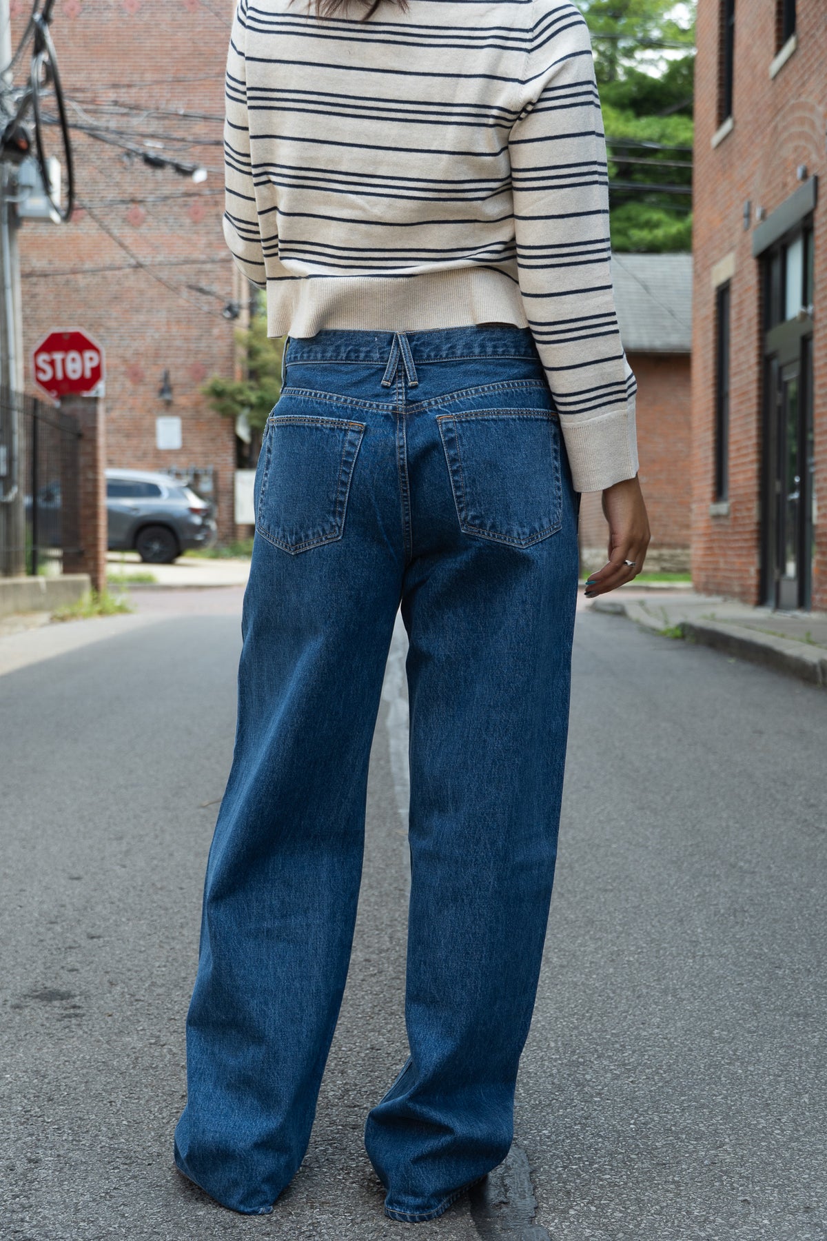 A person standing on a city street viewed from behind, wearing wide-leg blue denim jeans and a cream striped long-sleeve top.