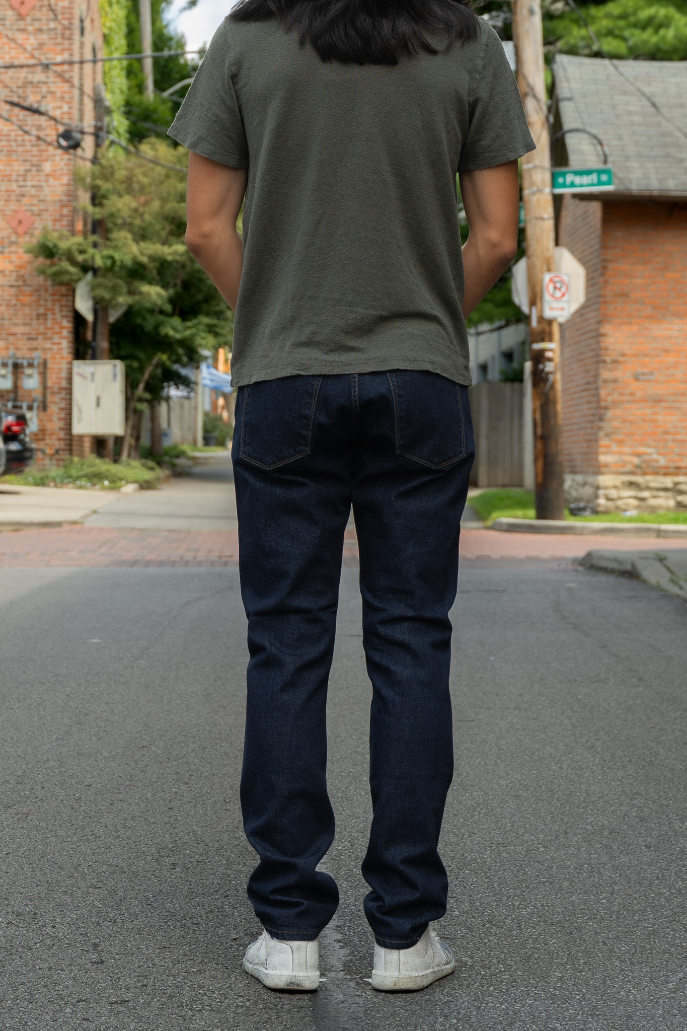 A person standing on a street, viewed from behind, wearing a dark olive green t-shirt, dark blue slim-fit jeans, and white sneakers.