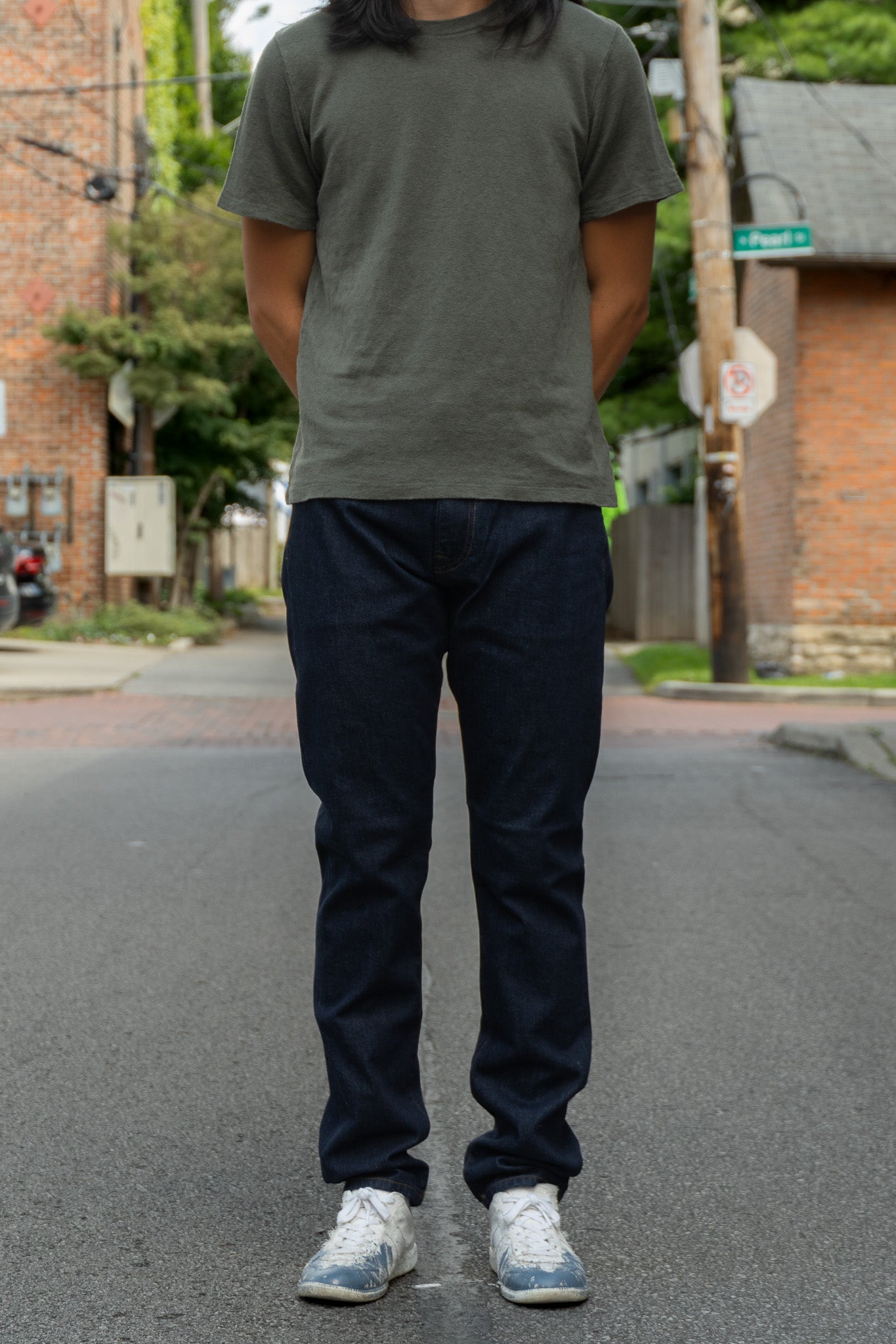 A person standing on an outdoor street wearing a dark olive green t-shirt, dark navy blue jeans, and white sneakers.