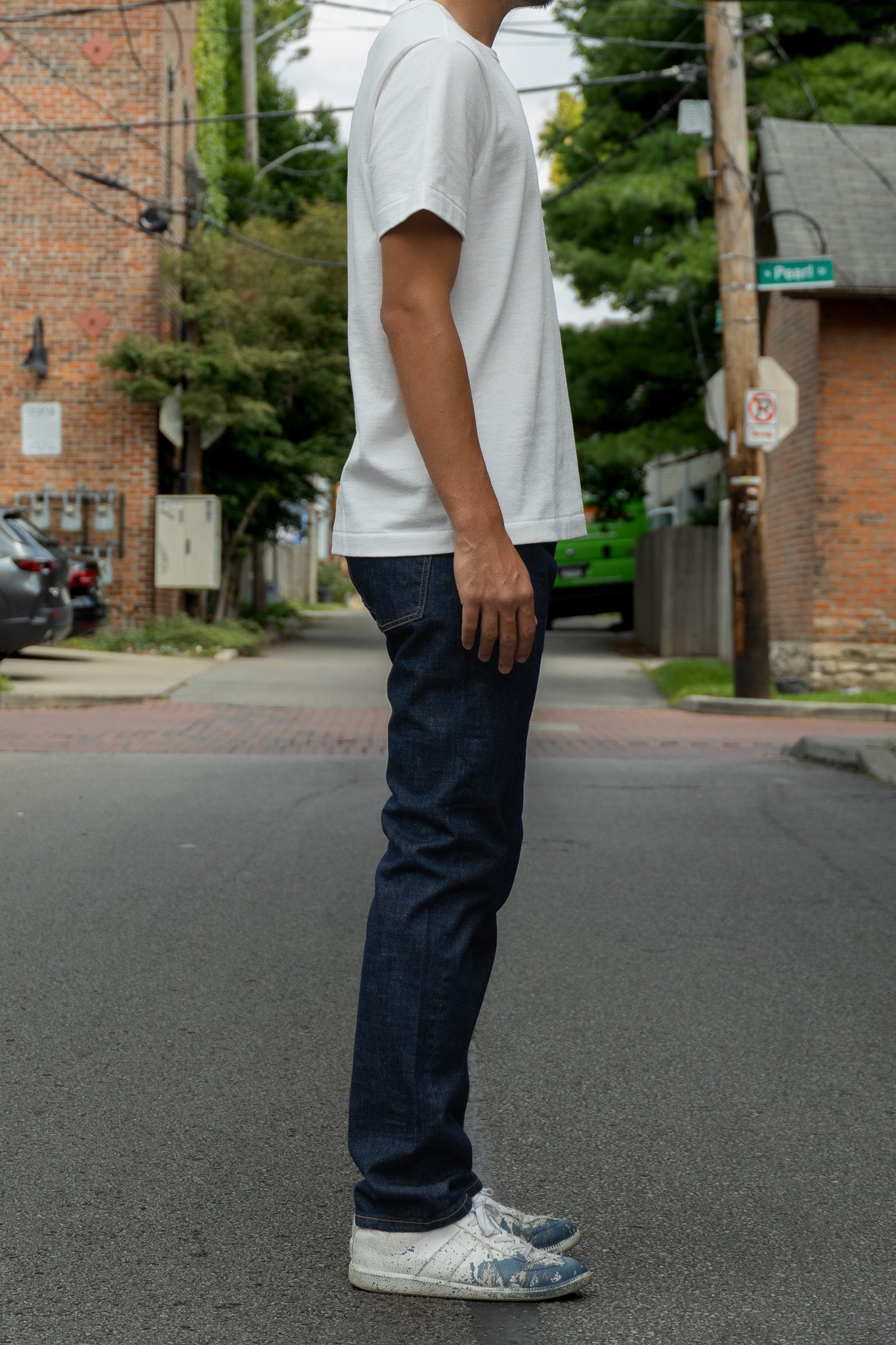 A person standing on a city street wearing a plain white short-sleeve T-shirt, dark blue jeans, and white sneakers