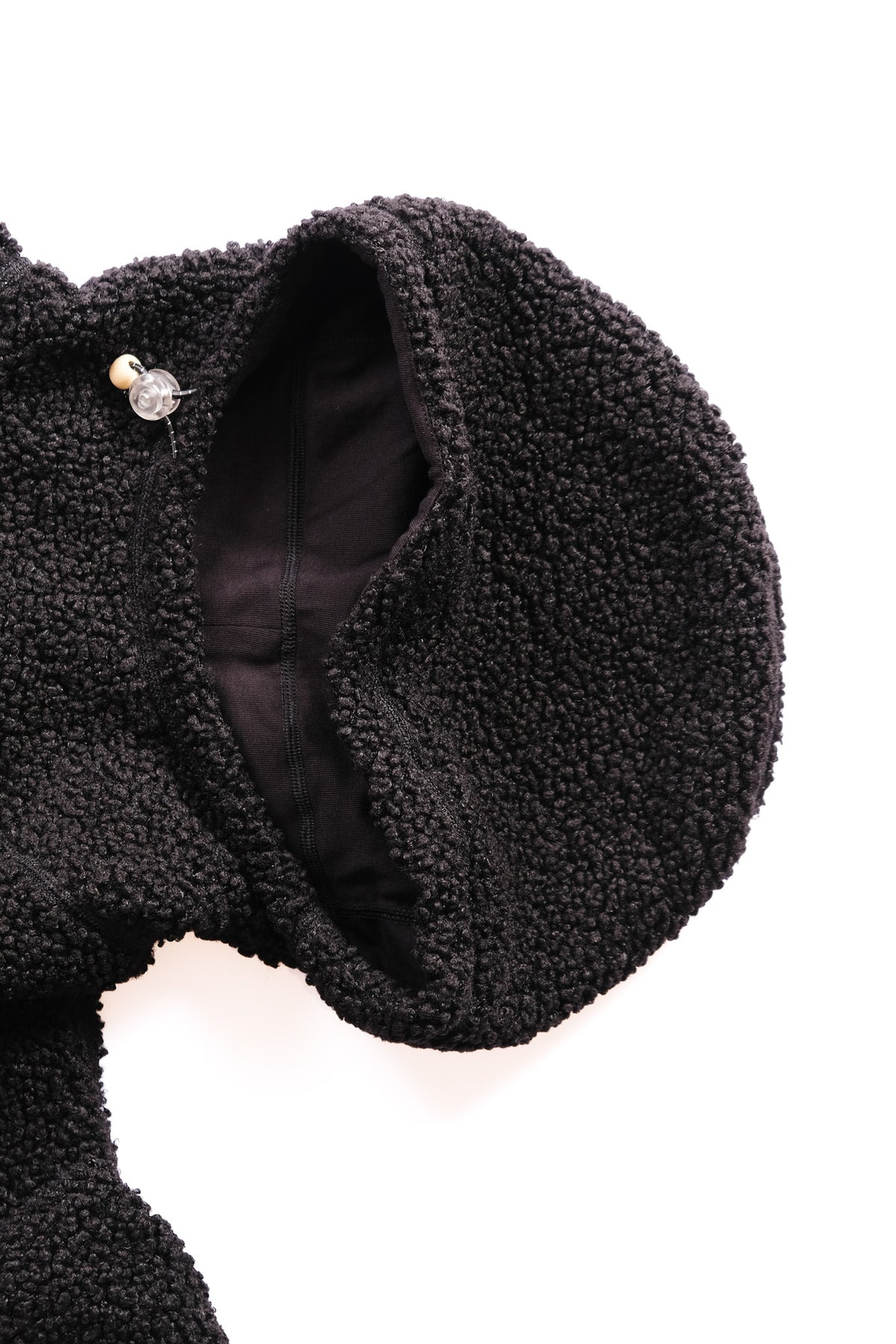 Close-up of a black sherpa fleece hood, showing the textured fabric, a black interior lining, and a small decorative toggle detail on a plain white background.