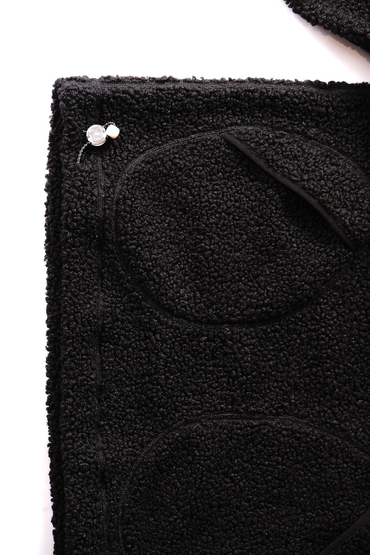 Close-up of a black textured sherpa fleece fabric item with a small white bead toggle along the hem  on a white background.