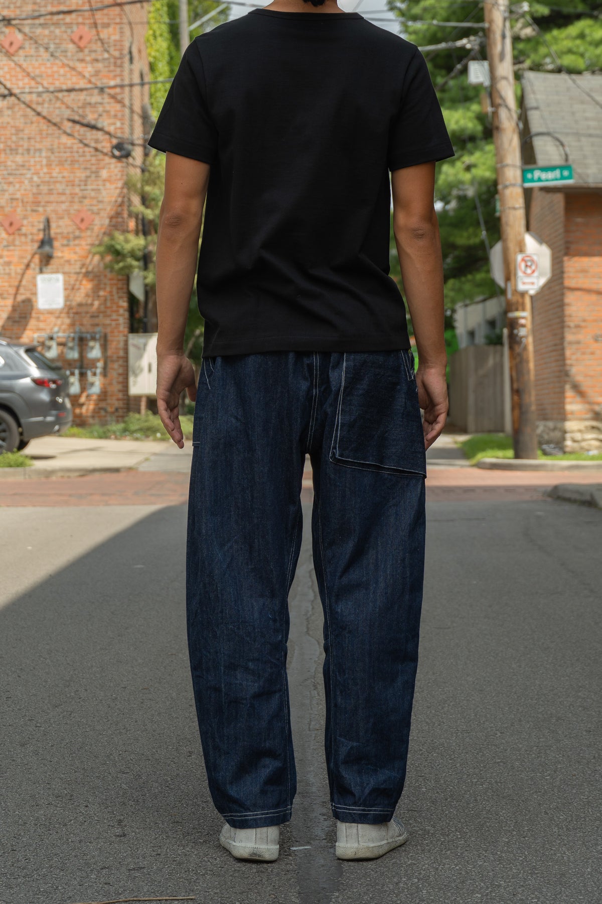 A person standing on a city street, viewed from behind, wearing loose dark blue denim jeans with a large back pocket, a black short-sleeve T-shirt, and light-colored sneakers.