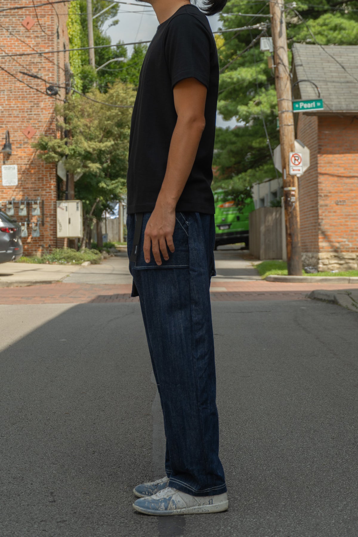 A person standing on an outdoor street wearing dark blue denim jeans, a black t-shirt, and light-colored sneakers, shown in profile.