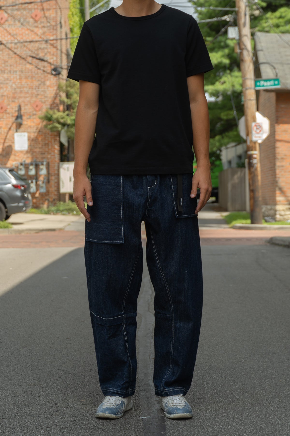 A person standing on a city street wearing a plain black short-sleeve T-shirt, dark blue wide-leg denim jeans with large patch pockets, and light blue sneakers.