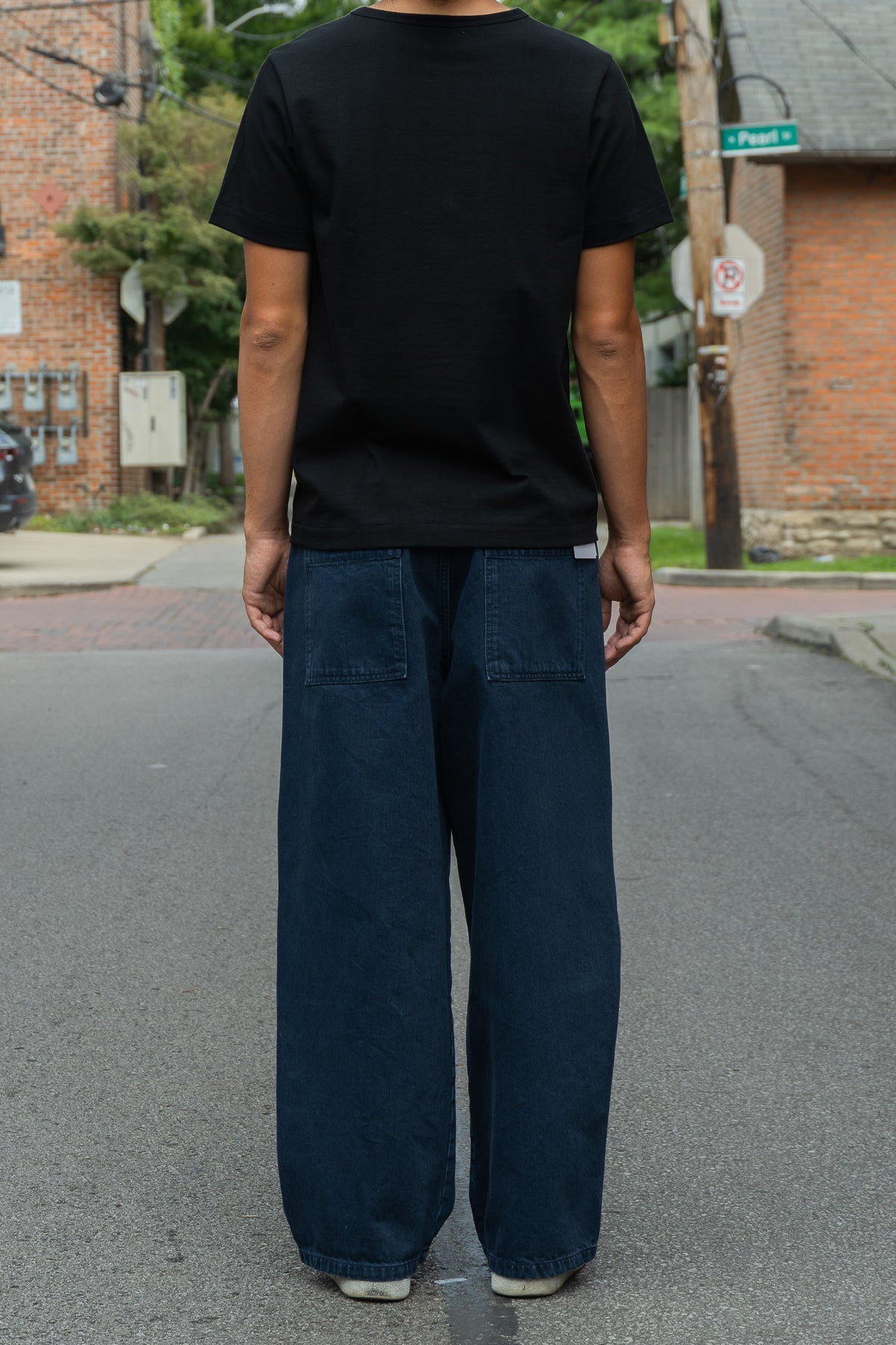 A person standing on a street, viewed from behind, wearing a black short-sleeve t-shirt and wide-leg dark blue jeans with back patch pockets.