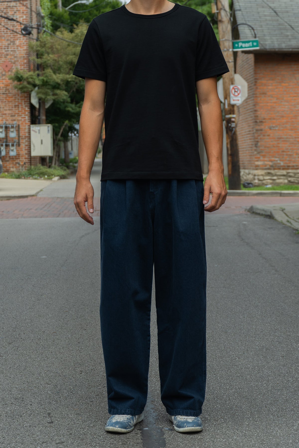 A person standing on an outdoor street wearing a plain black short-sleeve crewneck t-shirt and loose dark navy pants, with gray sneakers. 