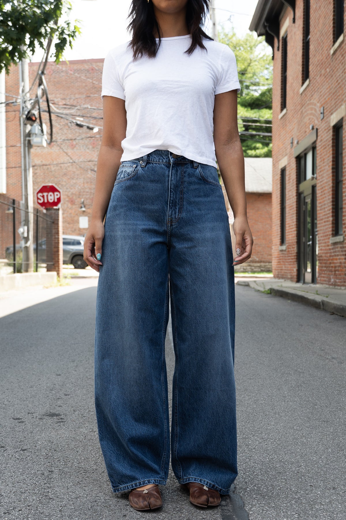 A person standing in the street wearing loose-fit medium blue wide-leg denim jeans, a white short-sleeve T-shirt, and brown flat shoes.