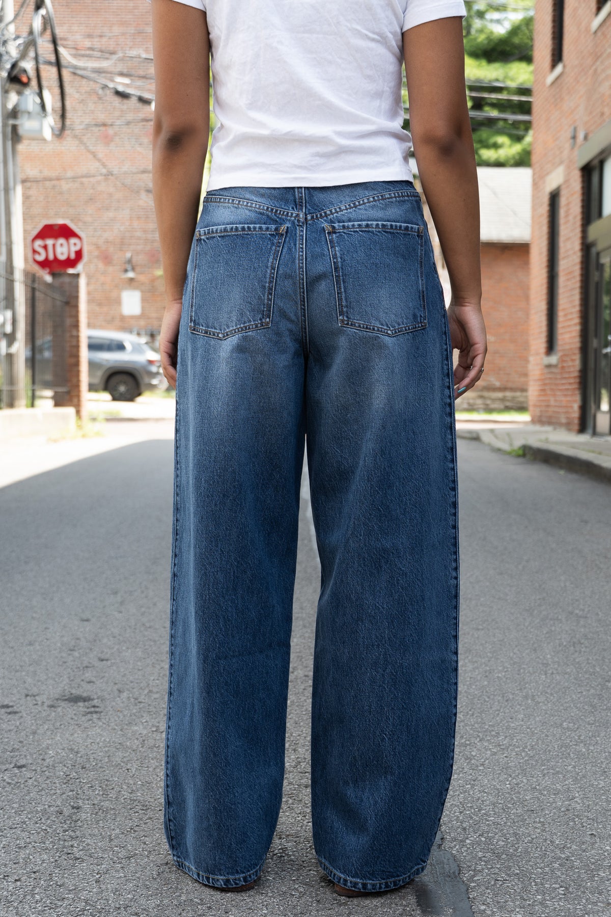 A person standing in the street, viewed from the back, wearing loose, wide-leg blue denim jeans and a white T-shirt.