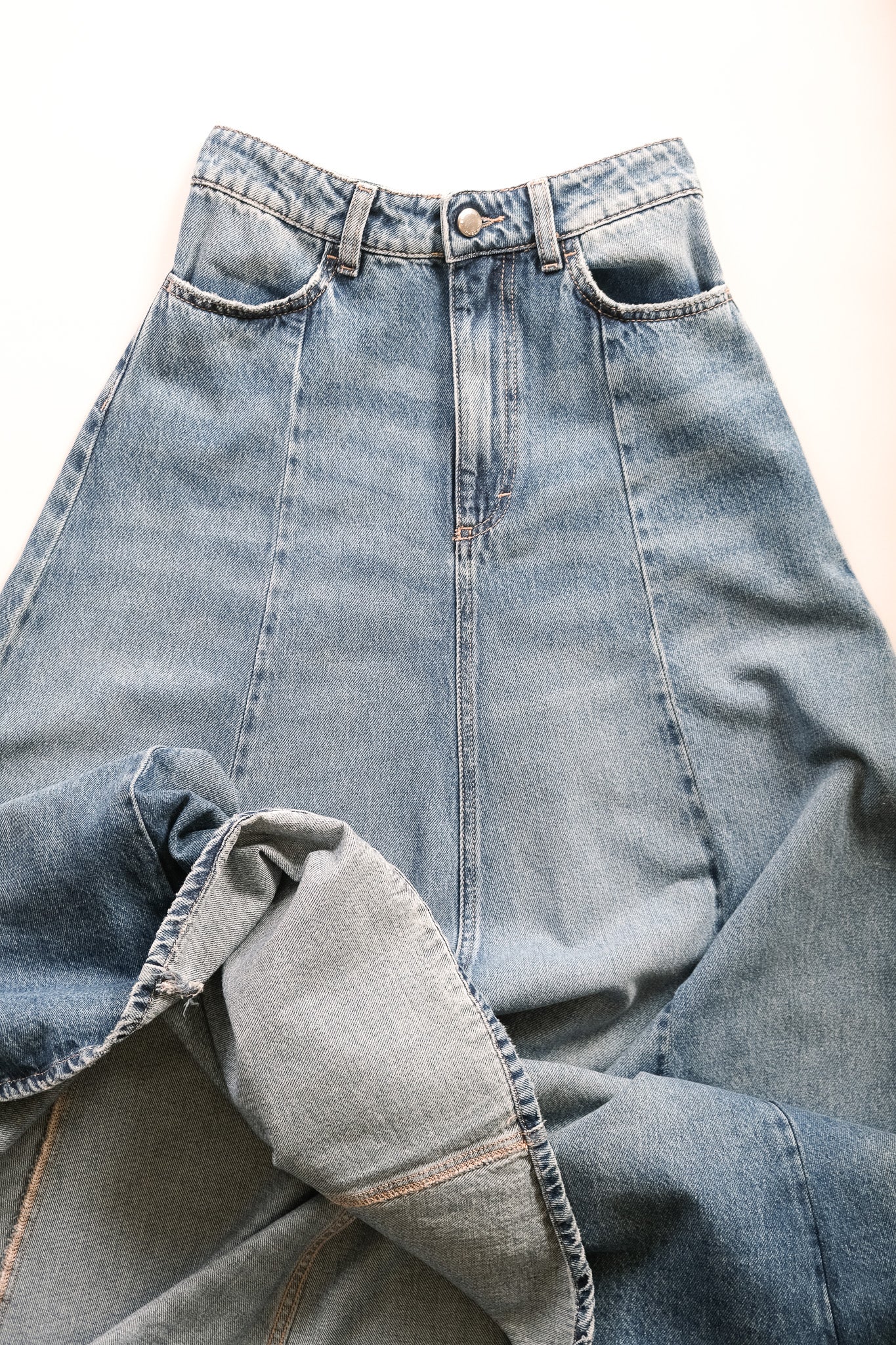 A close-up of a light blue denim skirt laid flat on a white background, showing the waistband, front pockets, and a folded section of the fabric.