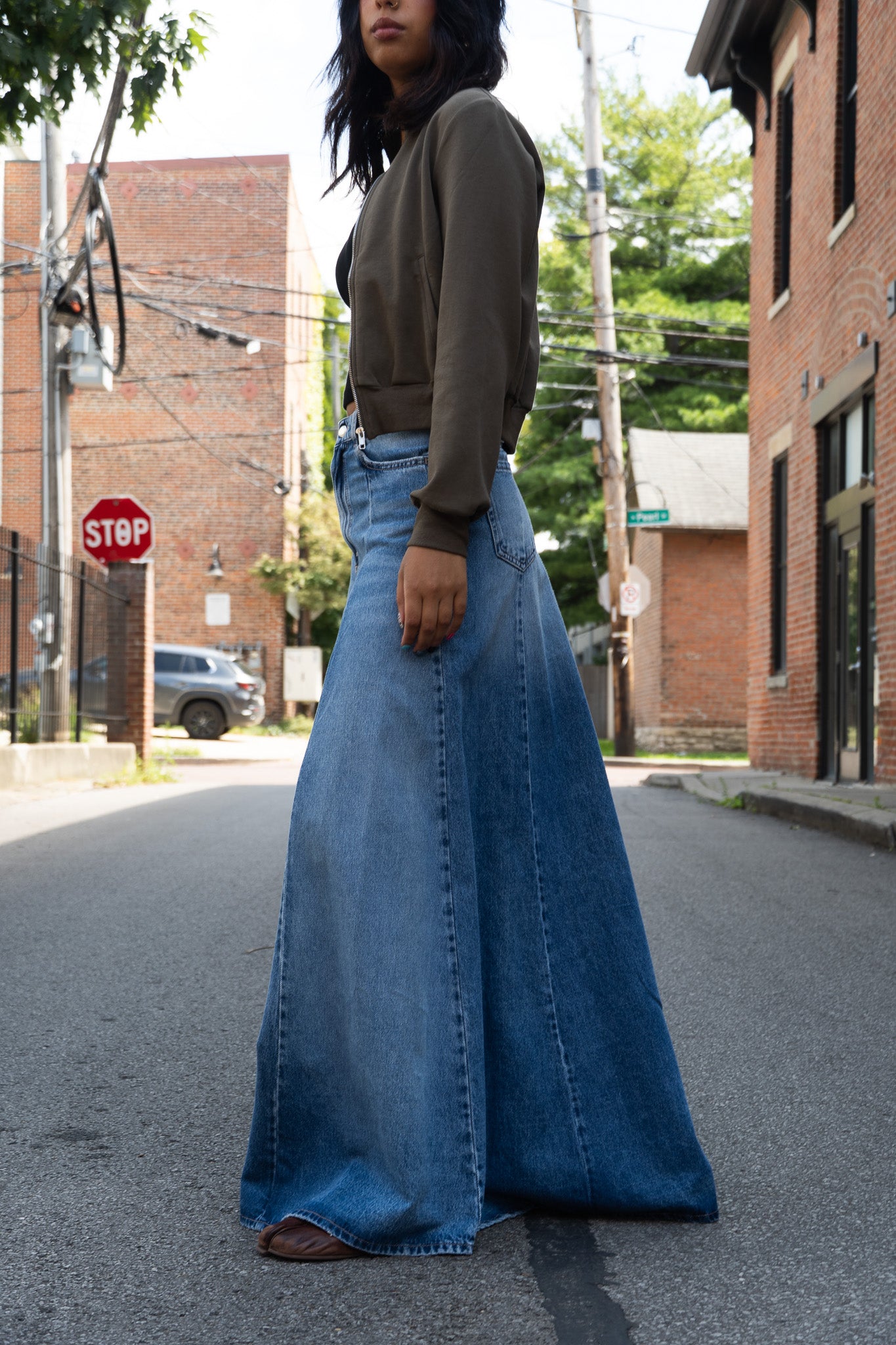 A person wearing a wide, floor-length blue denim skirt and a dark olive green zip jacket, standing in the street.