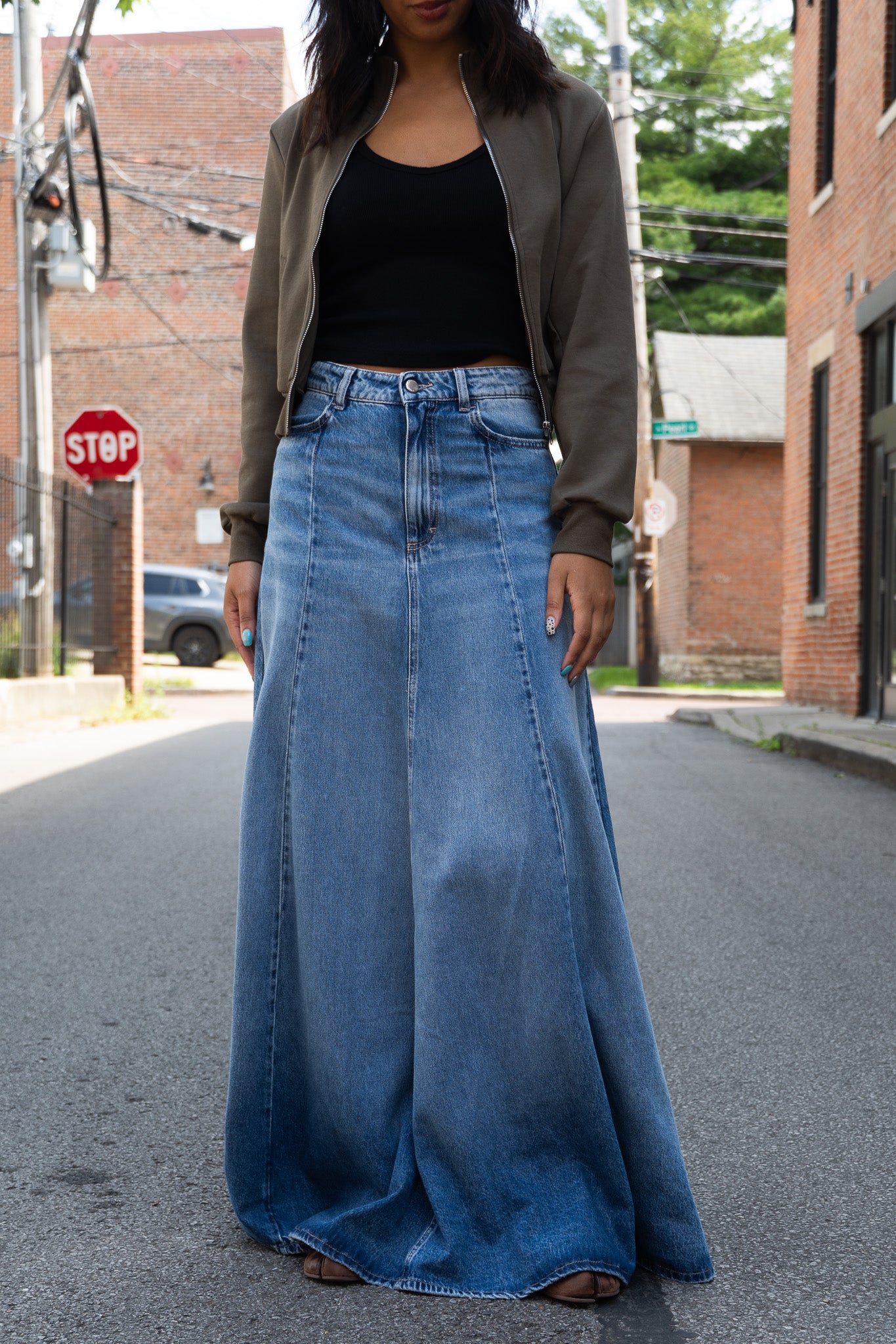 A woman standing on a city street wearing a long, wide blue denim skirt with a black tank top and an olive zip jacket.