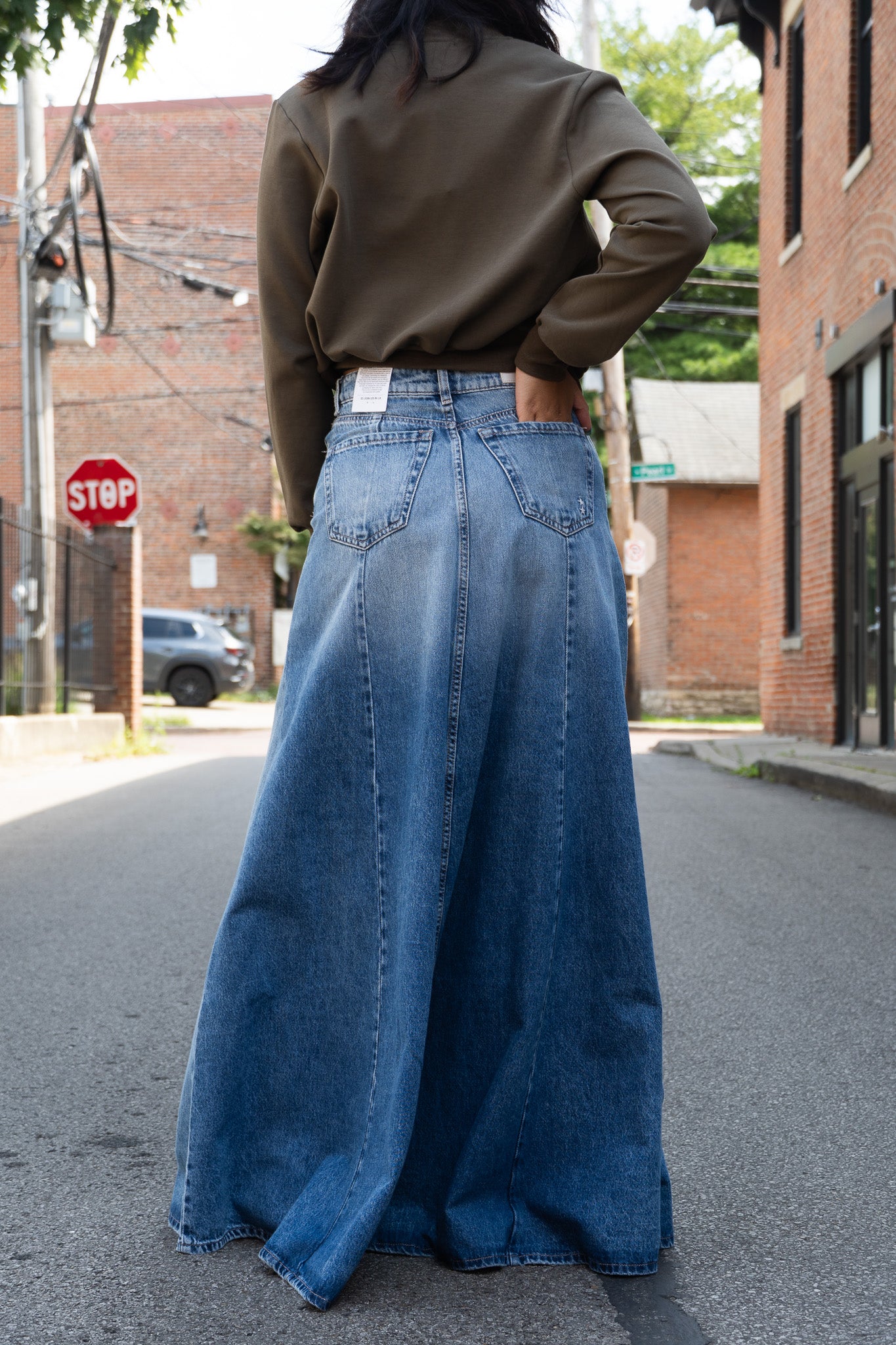 A person standing in the street wearing a long, wide-leg blue denim skirt with a  olive zip photographed from behind.