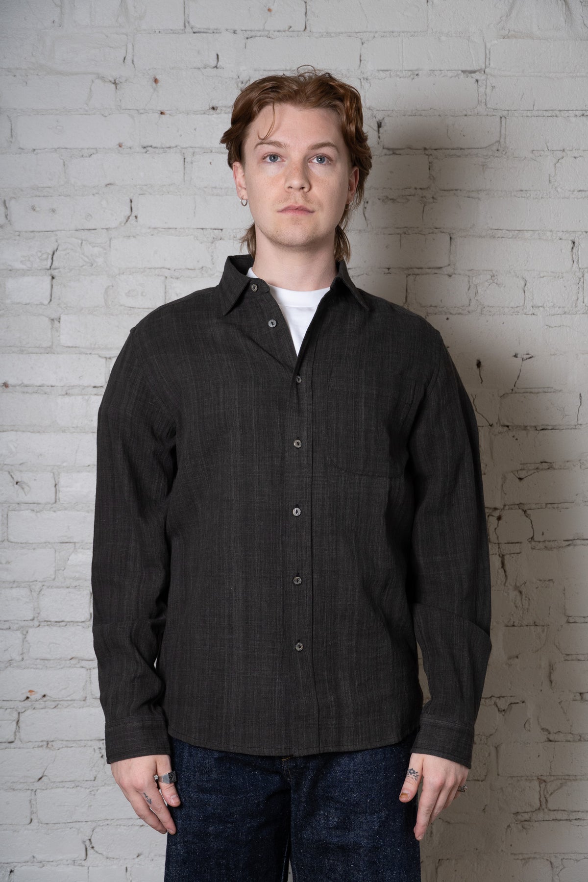 A man wearing a dark charcoal gray long-sleeve button-up shirt over a white undershirt and blue jeans, standing against a white brick wall.