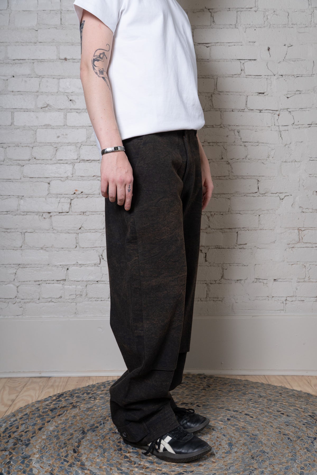 A person wearing loose, dark charcoal-brown wide-leg trousers with a white t-shirt and black sneakers, standing on a round woven rug against a white brick wall.