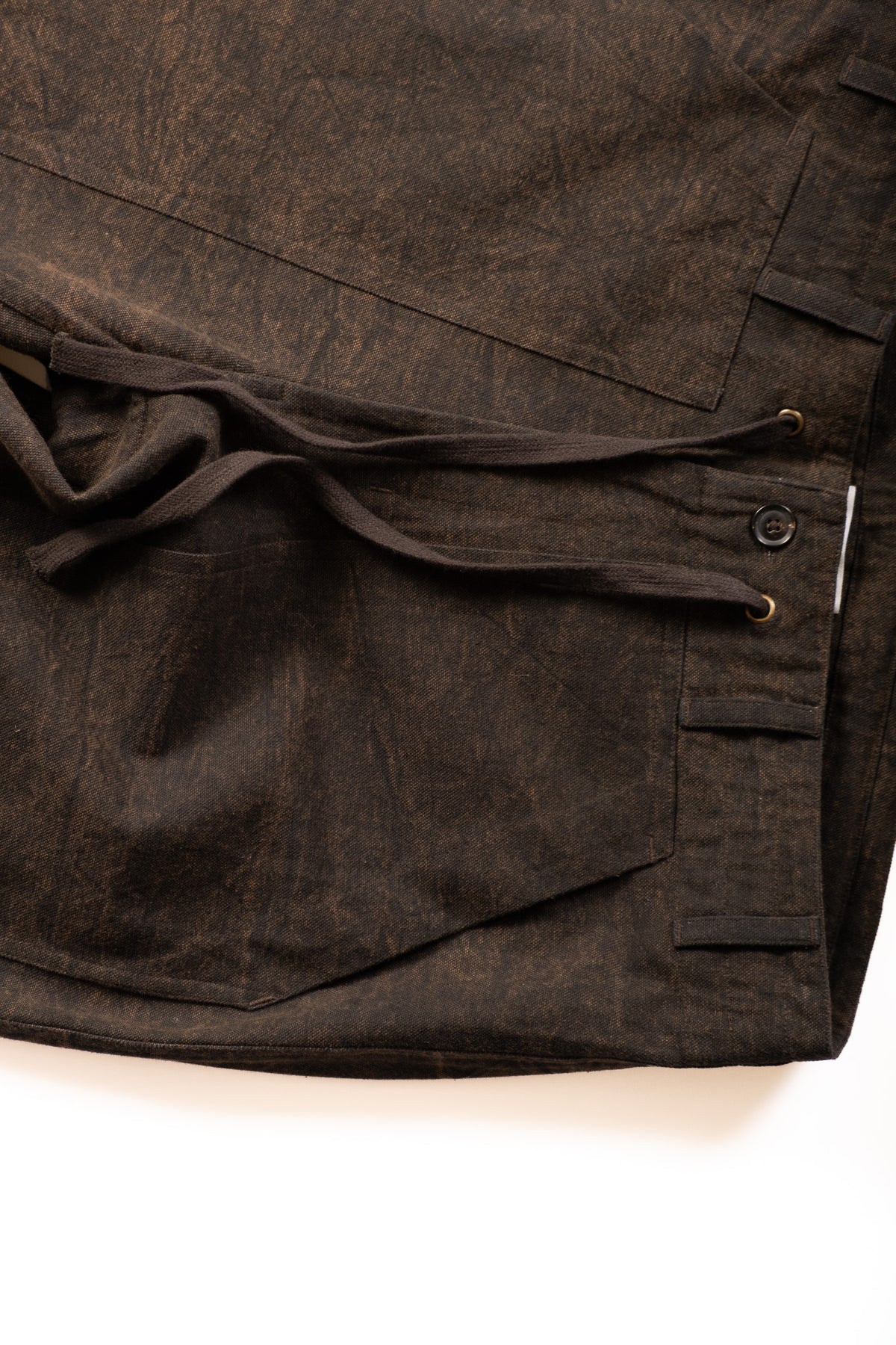 Close-up of a dark brown textured fabric garment, showing a drawstring waist, button closure, and side pocket on a white background.