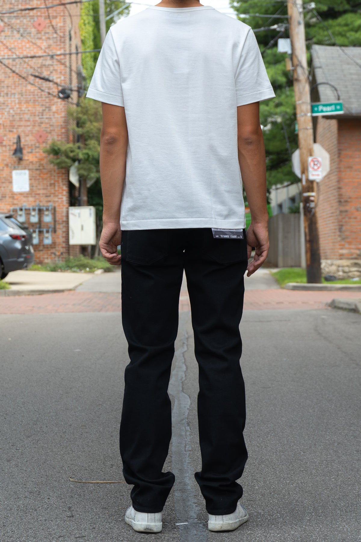 A person standing on a city street, viewed from behind, wearing a plain white short-sleeve T-shirt, black jeans, and white sneakers.