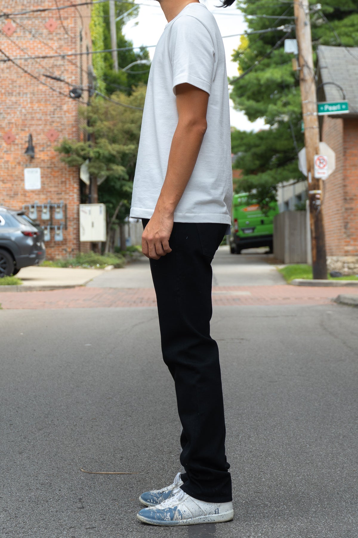 A person standing on an outdoor street wearing a white short-sleeve t-shirt, black jeans, and white sneakers viewed from the side.